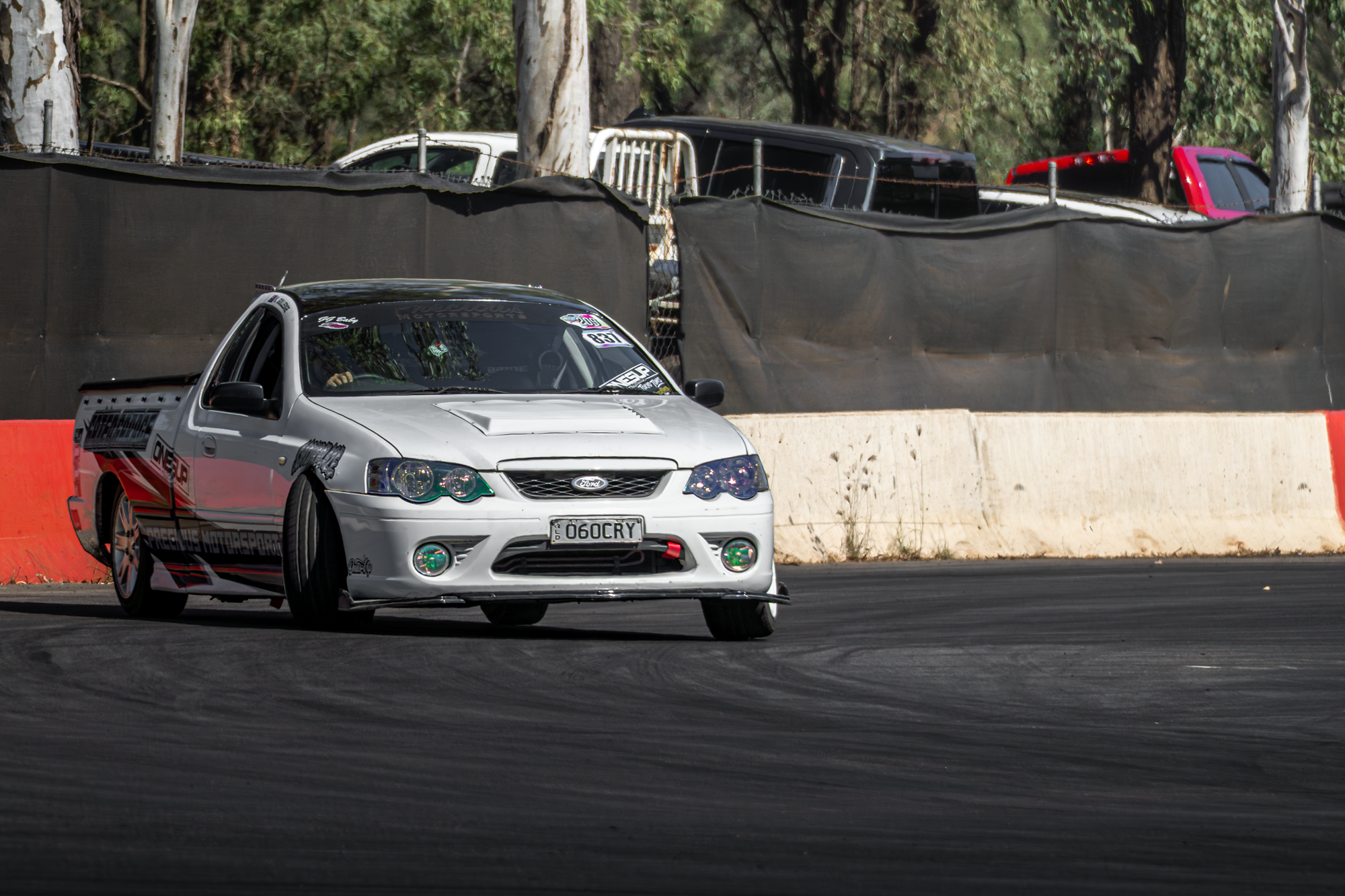 White racing car with black and red decals on a race track, taking a turn with a rocky and wooded background.