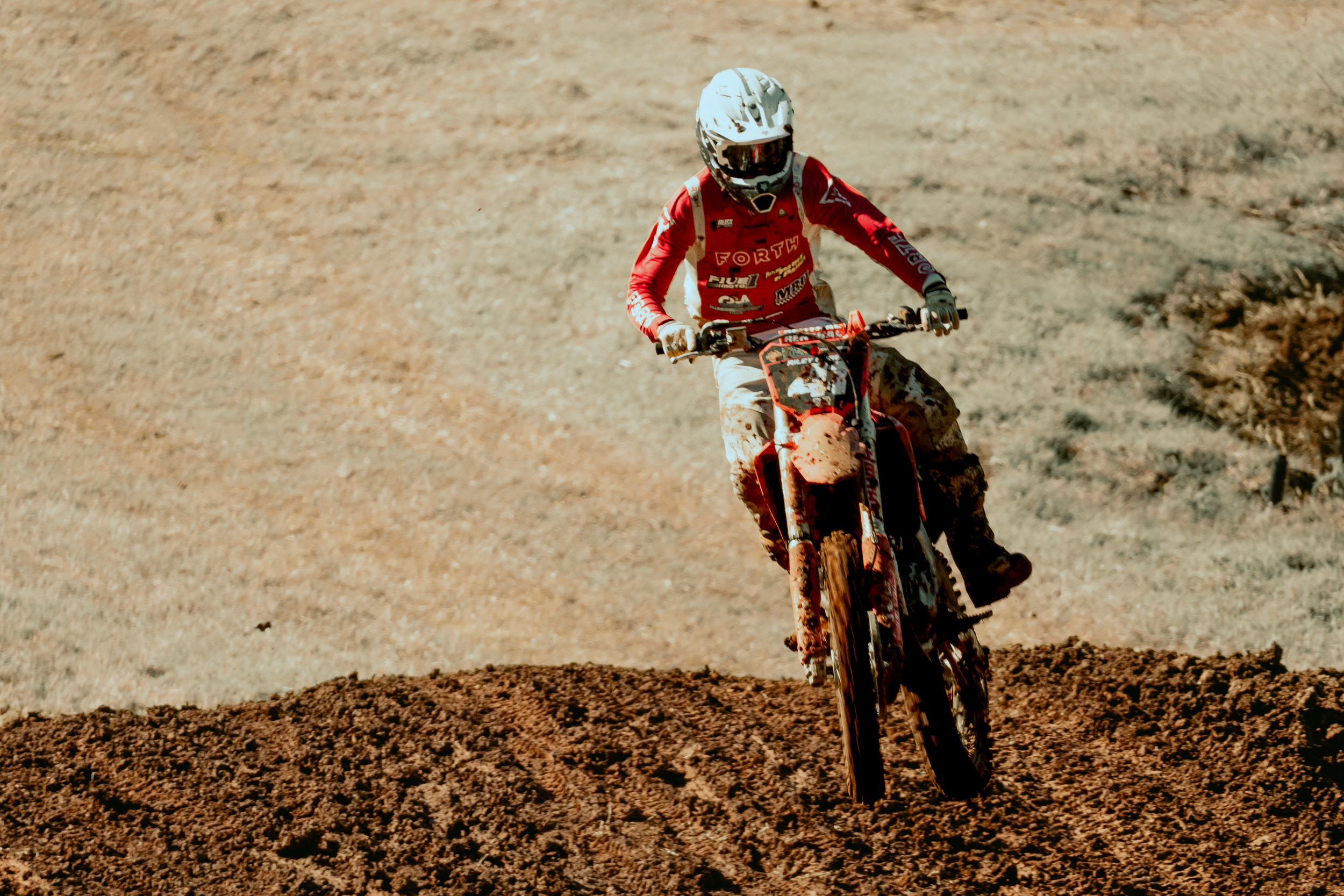 A person wearing a white helmet and red racing gear riding a dirt bike on a dusty off-road trail.