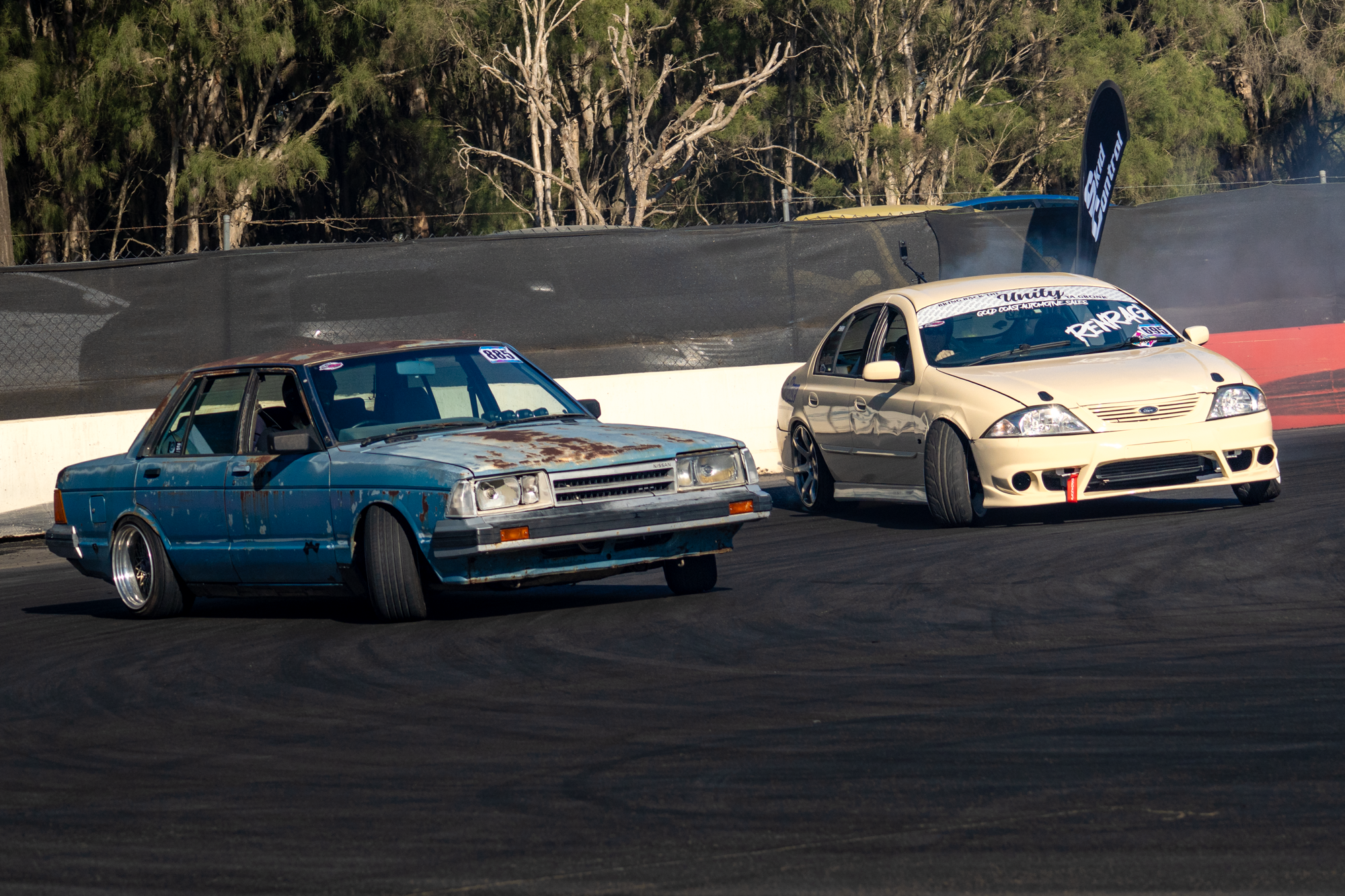 Two race cars on a track, one rusted blue with a boxy shape, and one cream-colored with a more modern aerodynamic design, with trees in the background.
