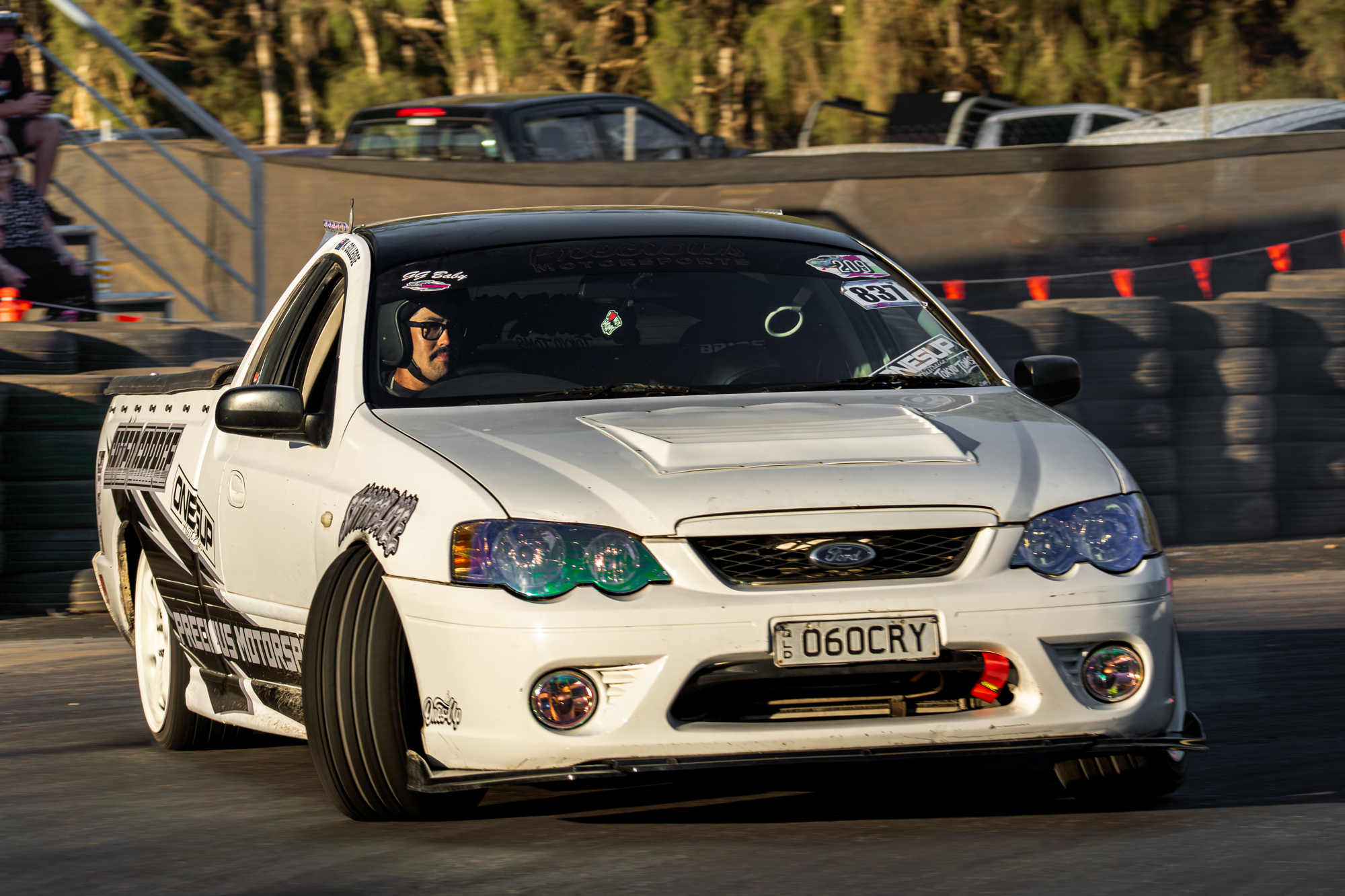 A white sports car with racing decals, driving on a race track with cars and trees in the background.
