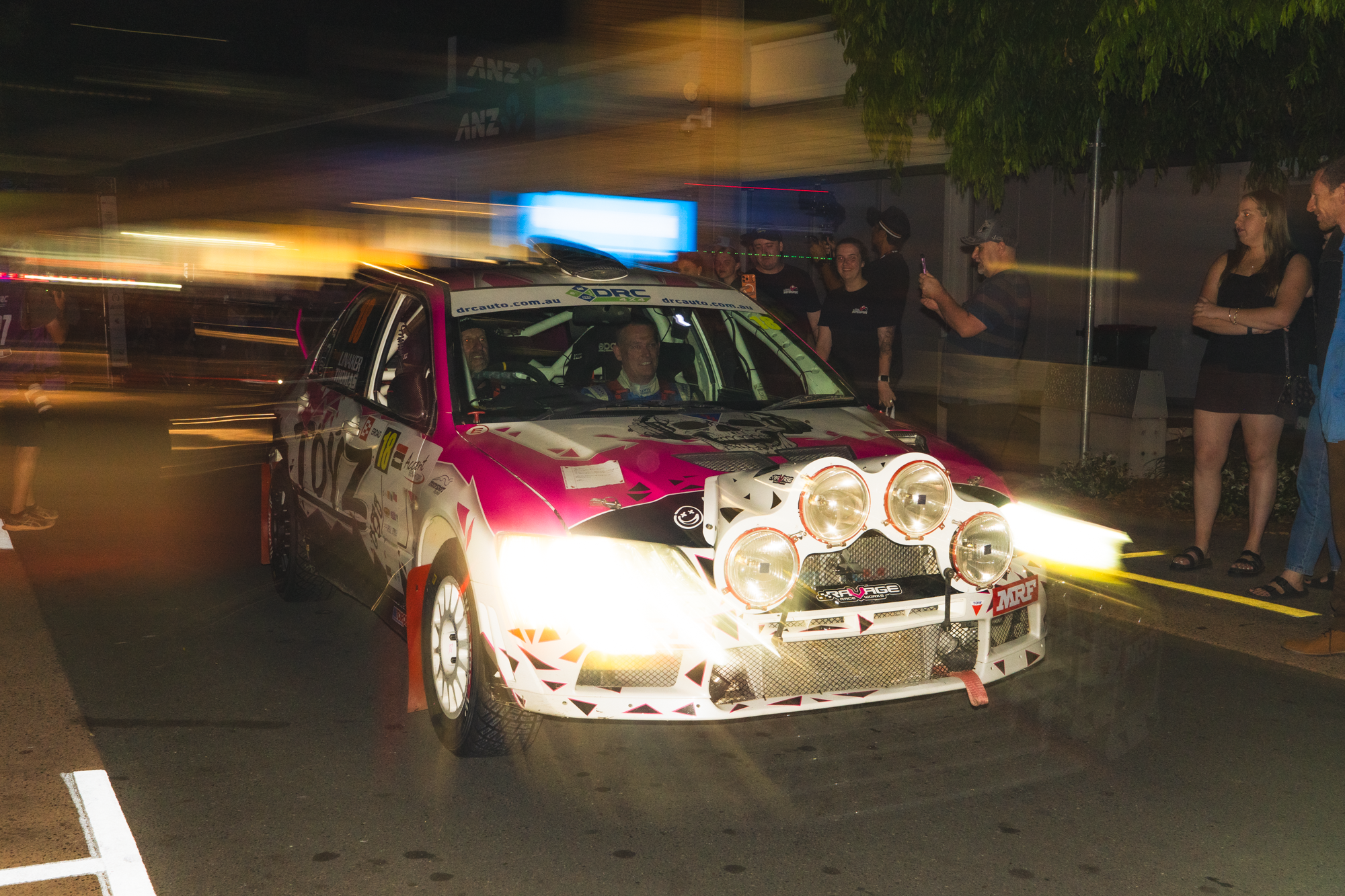 A rally car with pink, white, and black accents is parked on the street at night, illuminated by headlights. Several people are gathered around, some taking photos and others chatting, with blurred motion lights in the background.