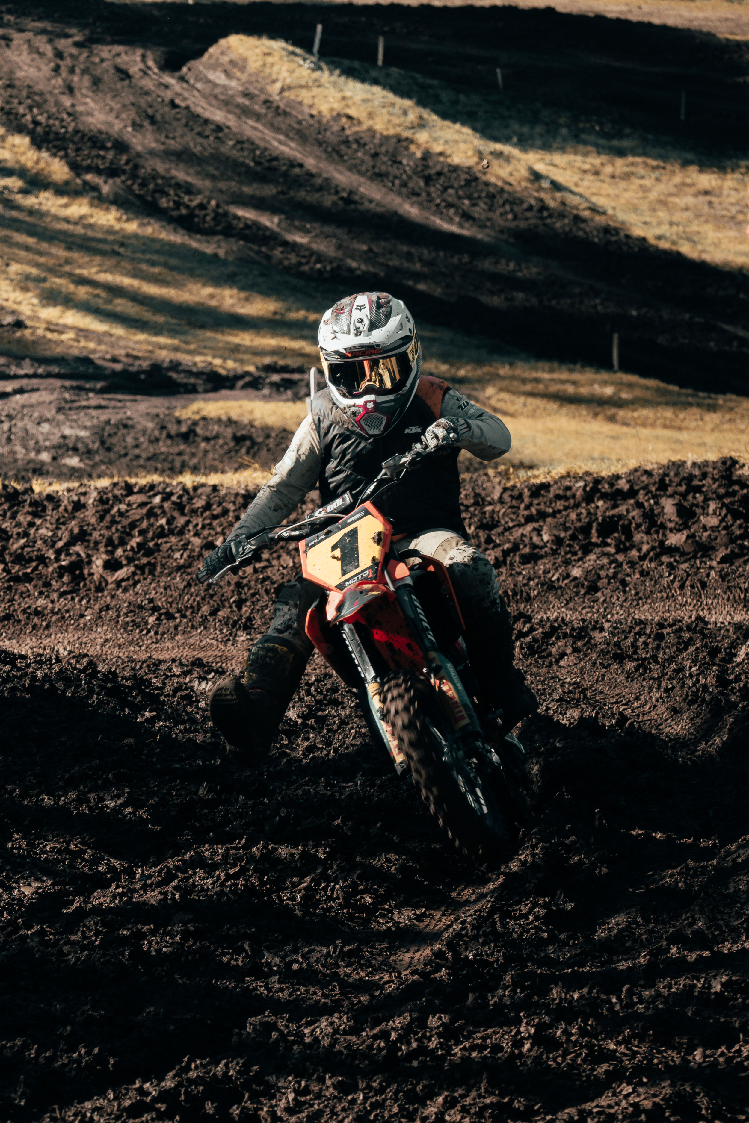 A motocross rider wearing a helmet and protective gear riding a dirt bike on a rugged, muddy track with a hilly landscape in the background.