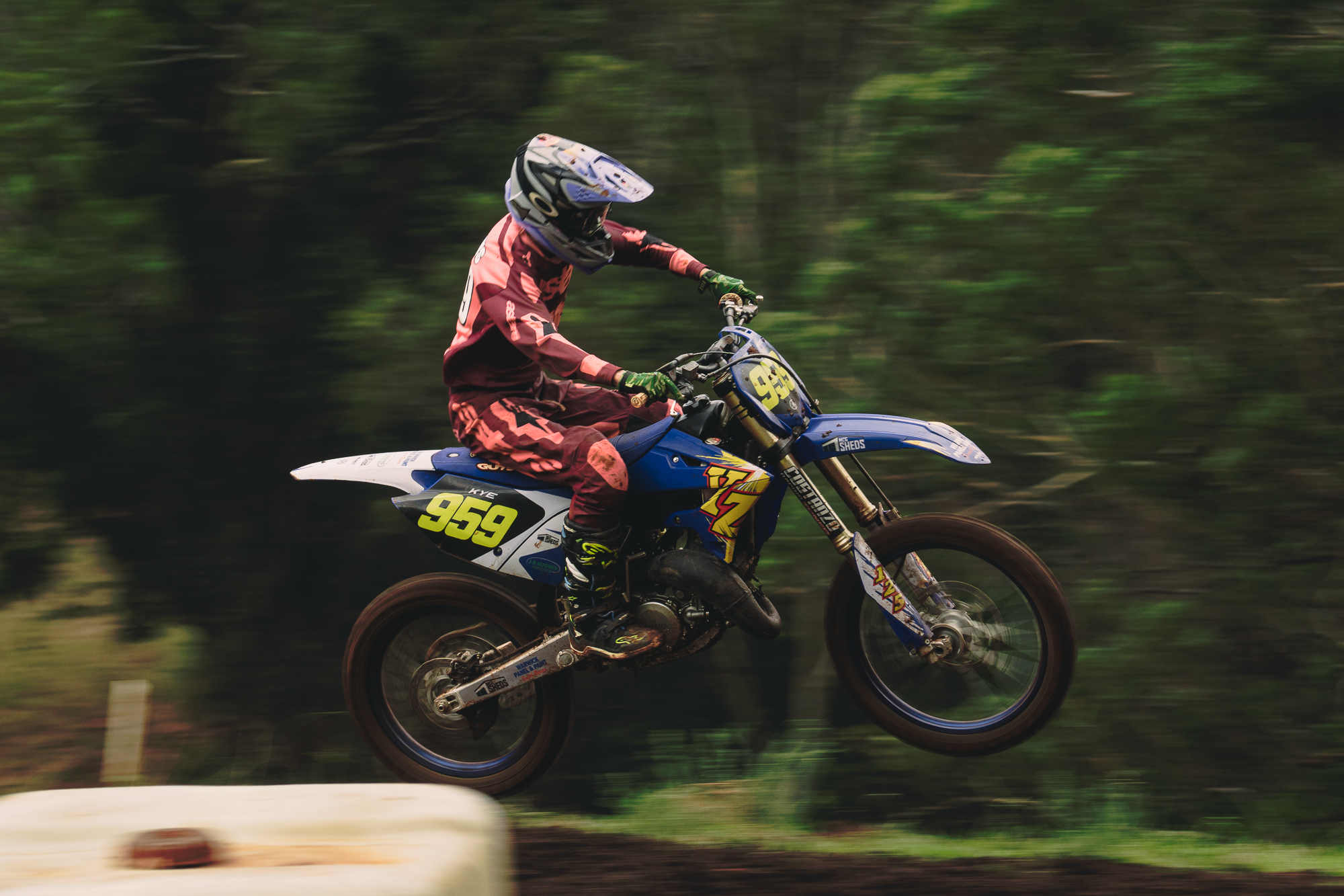 A motocross rider wearing a helmet and racing gear, riding a blue and white dirt bike numbered 959, speeding through a forested area with blurred greenery in the background.