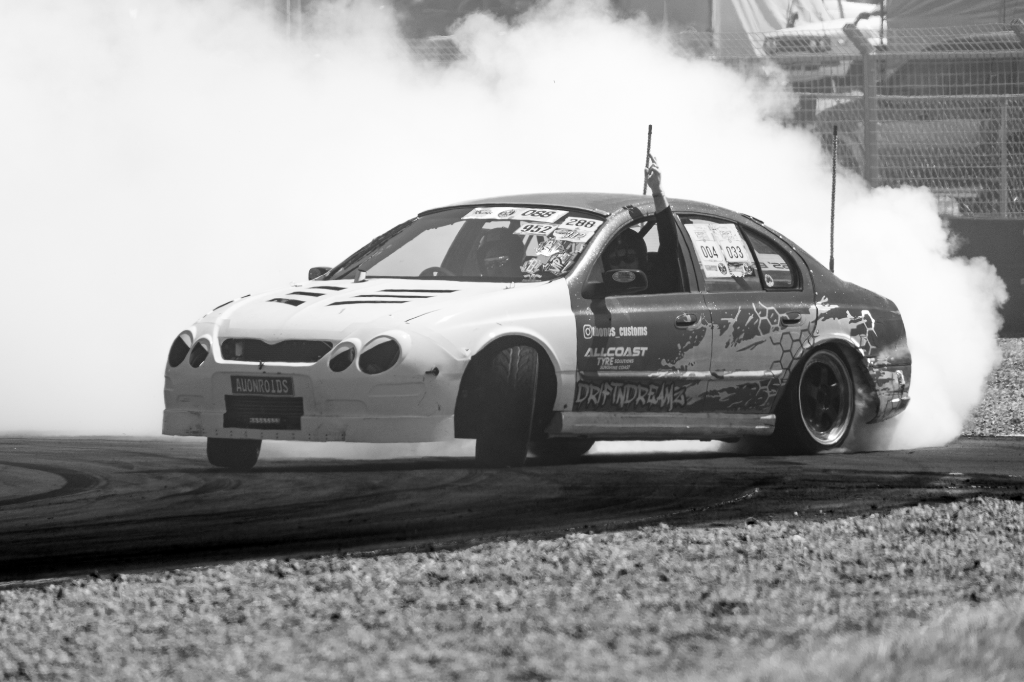 A race car performing a burnout during a motorsport event, with smoke billowing from the tires, and racing stickers visible on the car. The car is on a dirt track with a fence and vehicles in the background.