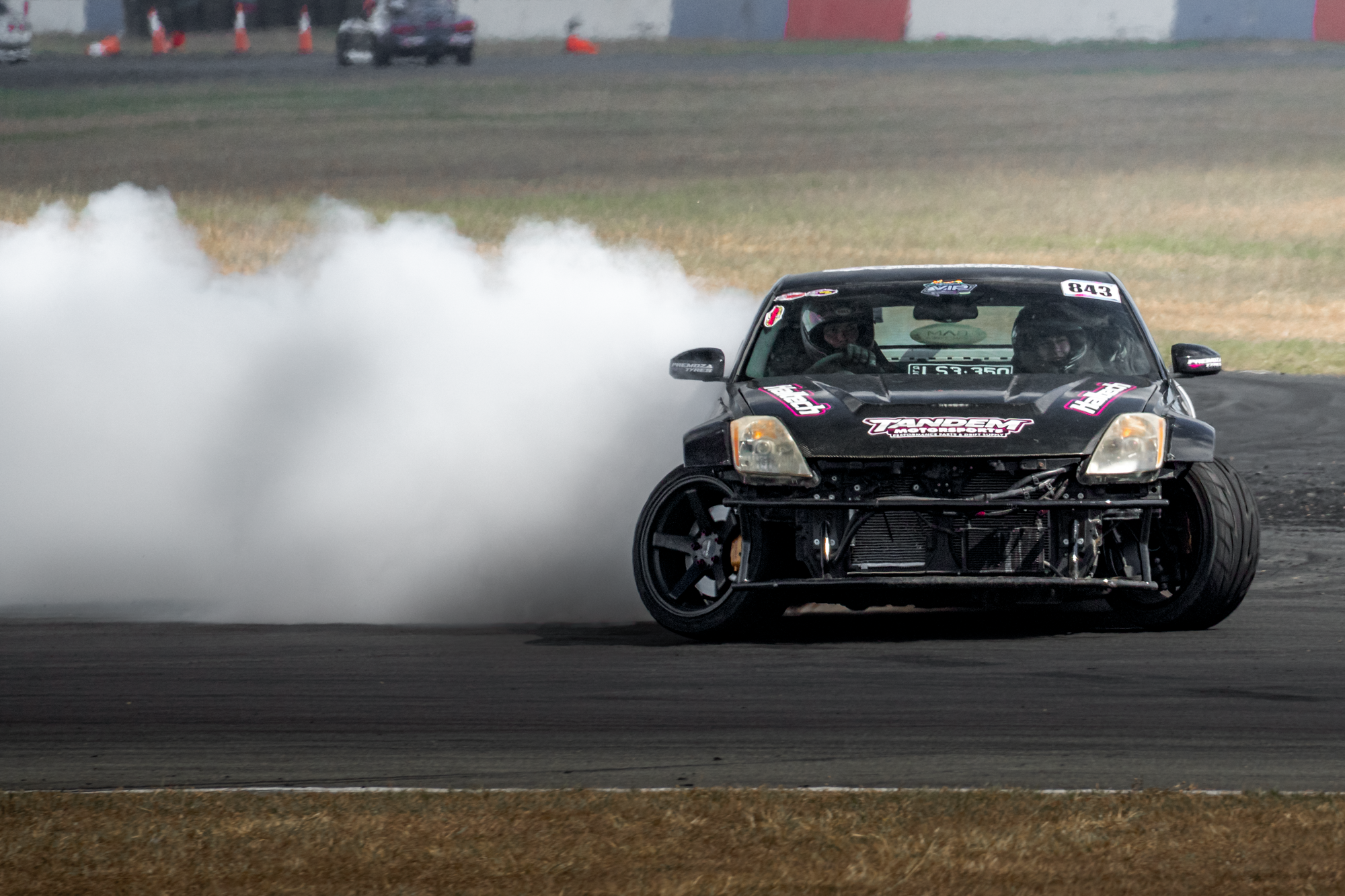 A black race car drifting on a racetrack with white smoke coming from the tires. The car has racing decals and the driver is wearing a helmet.