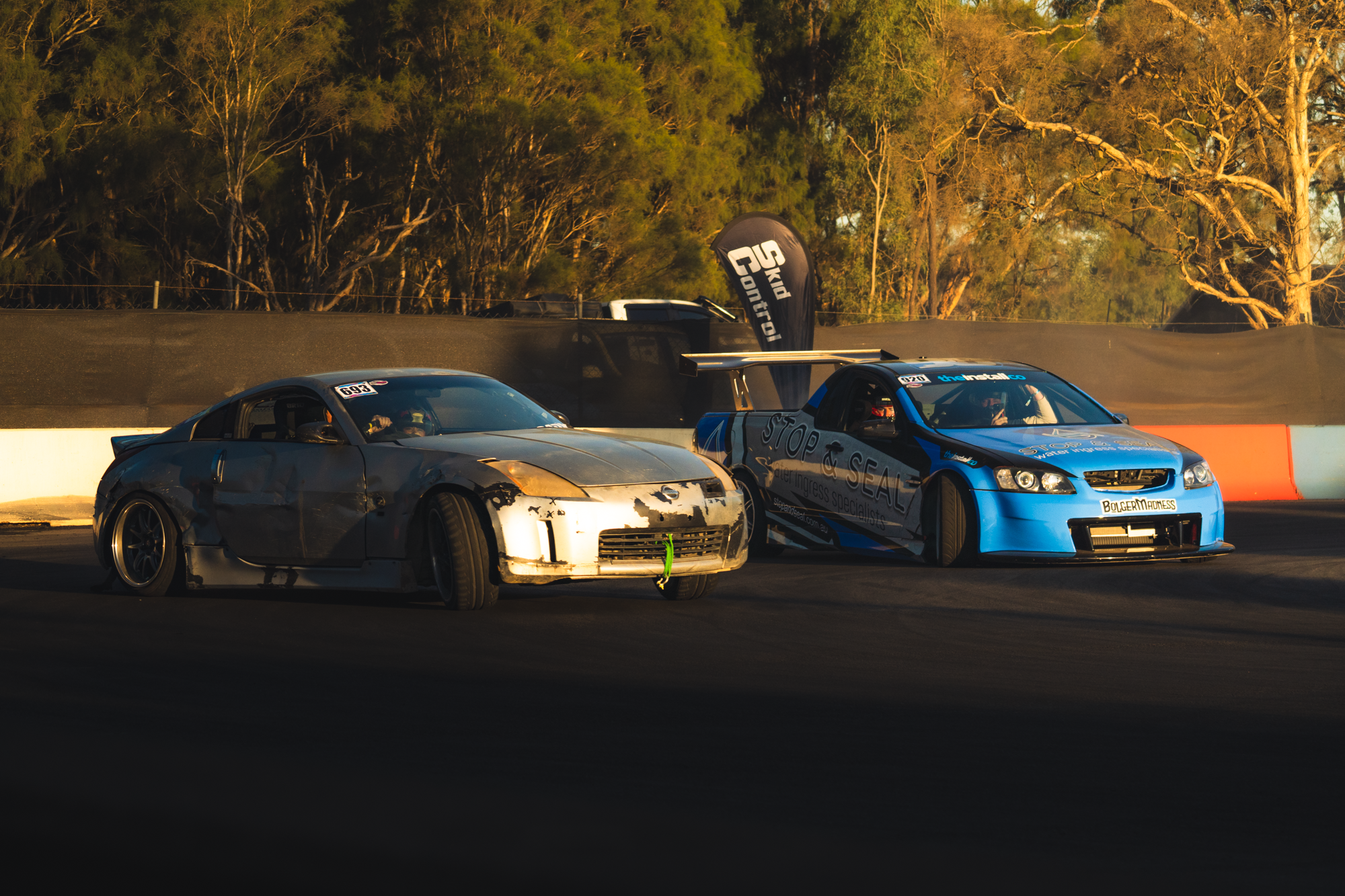 Two race cars on a track, one silver and one blue, with a background of trees and a black windscreen banner.