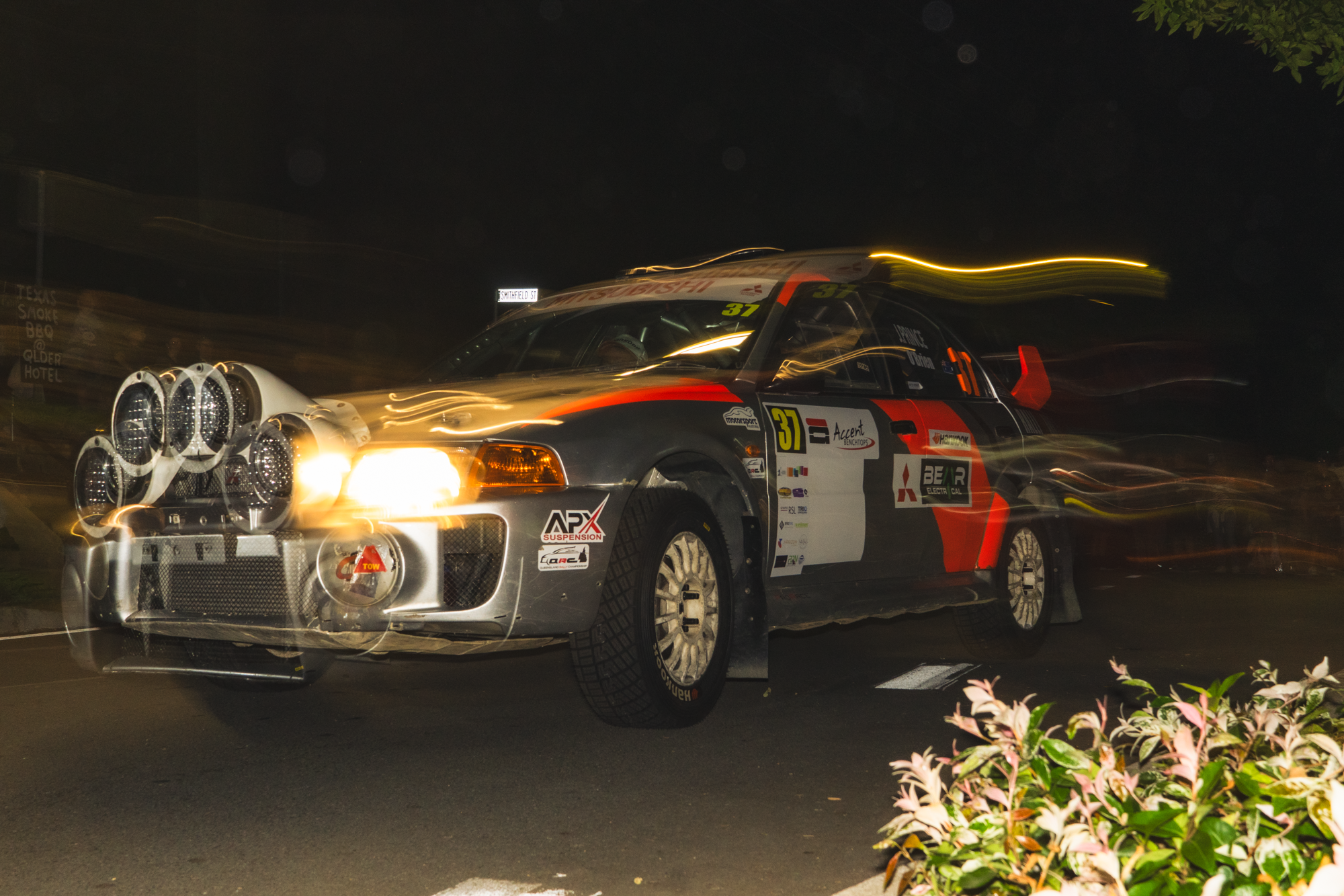 A rally car speeding at night, with light trails indicating motion, parked beside a road with some plants in the foreground and a sign with partially visible text in the background.