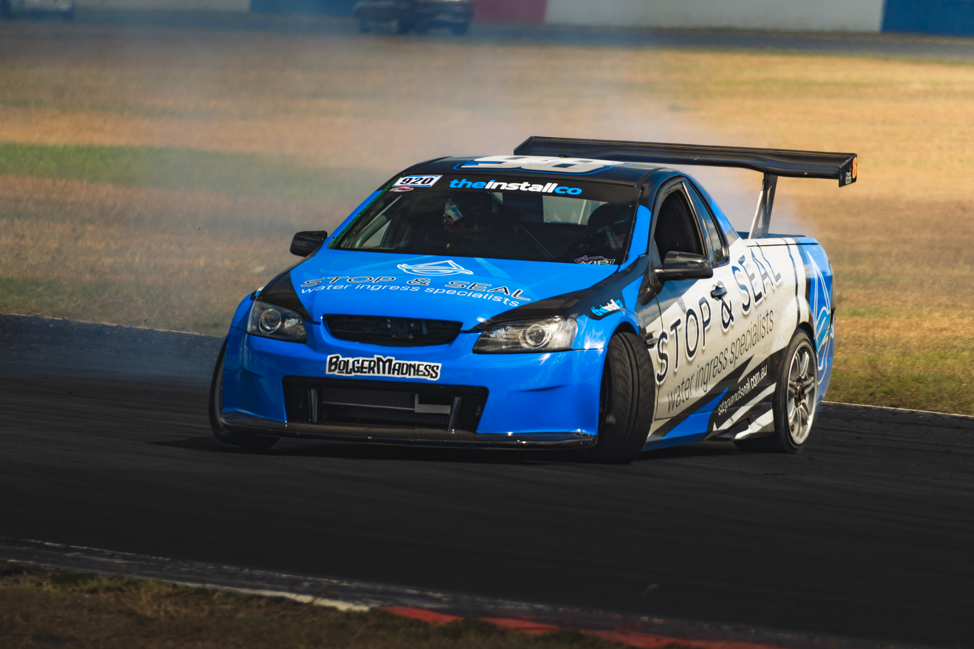 Blue race car with sponsor logos and a large rear wing speeding on a racetrack, kicking up dust in the background.