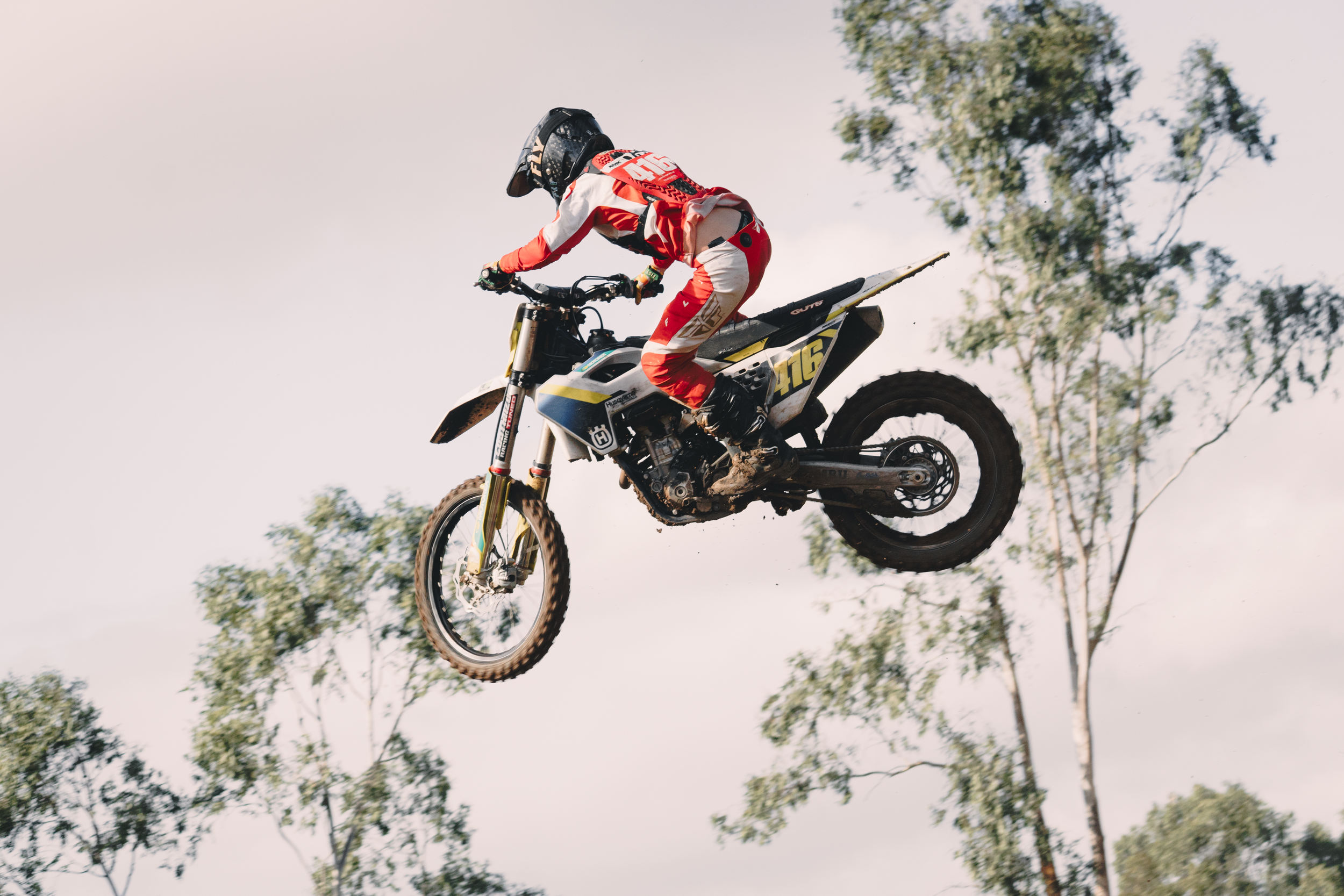 Motocross rider in red and white gear and black helmet jumping a dirt bike over a jump with trees in the background.