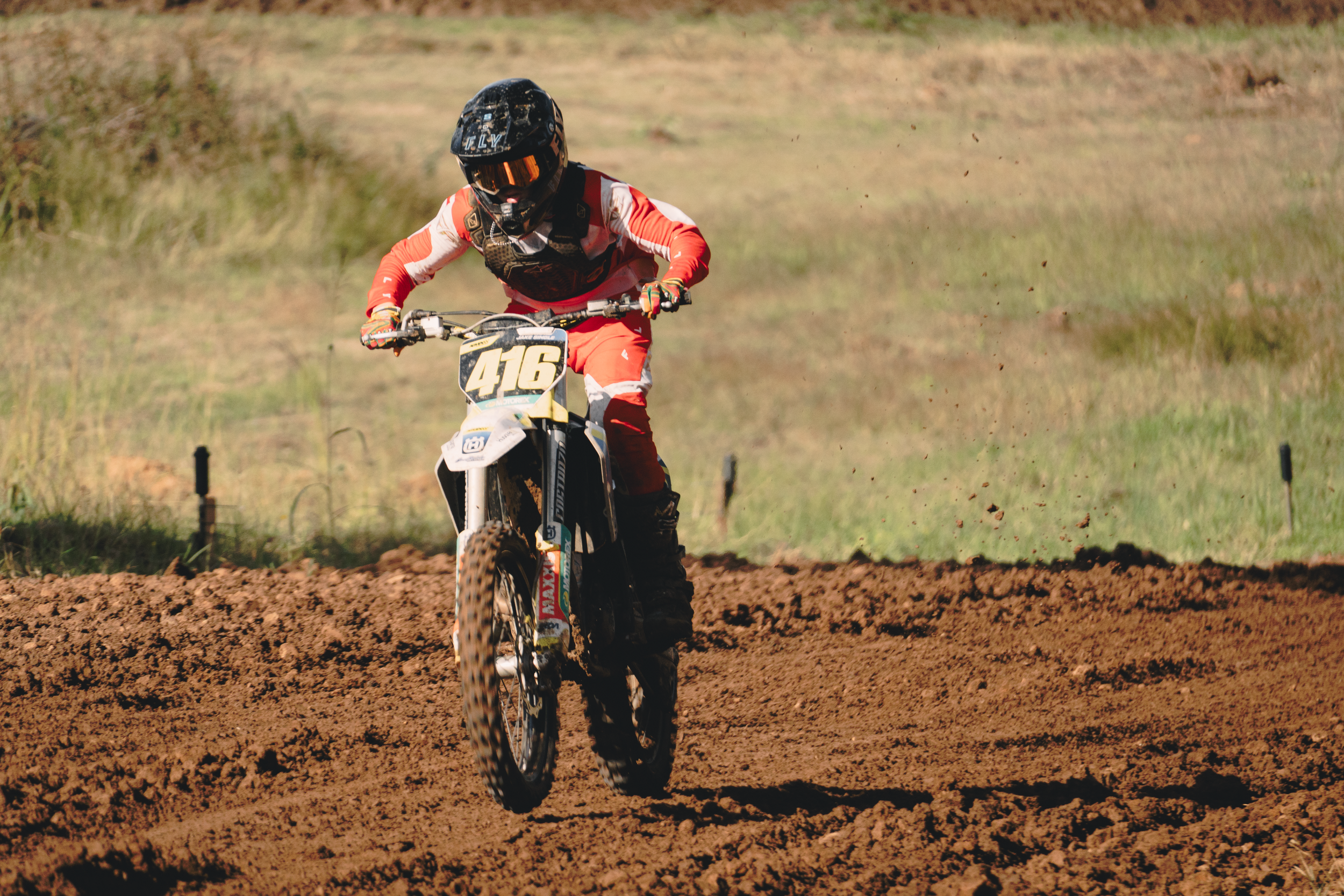 A person riding a dirt bike on a dirt track with an open landscape background, wearing a black helmet, orange goggles, red and white riding gear, and gloves, with the number 416 on the bike.