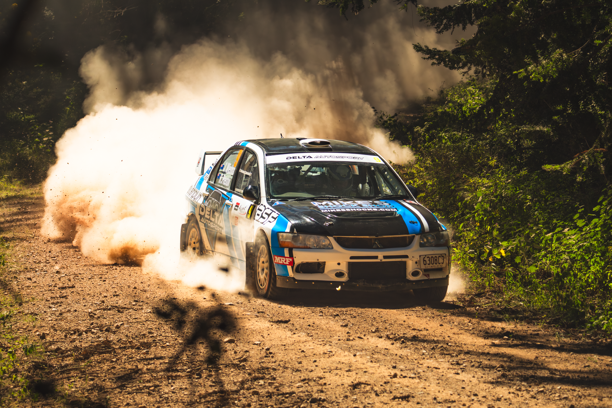 A rally car speeding on a dirt road, kicking up a large cloud of dust and debris while surrounded by trees and foliage.