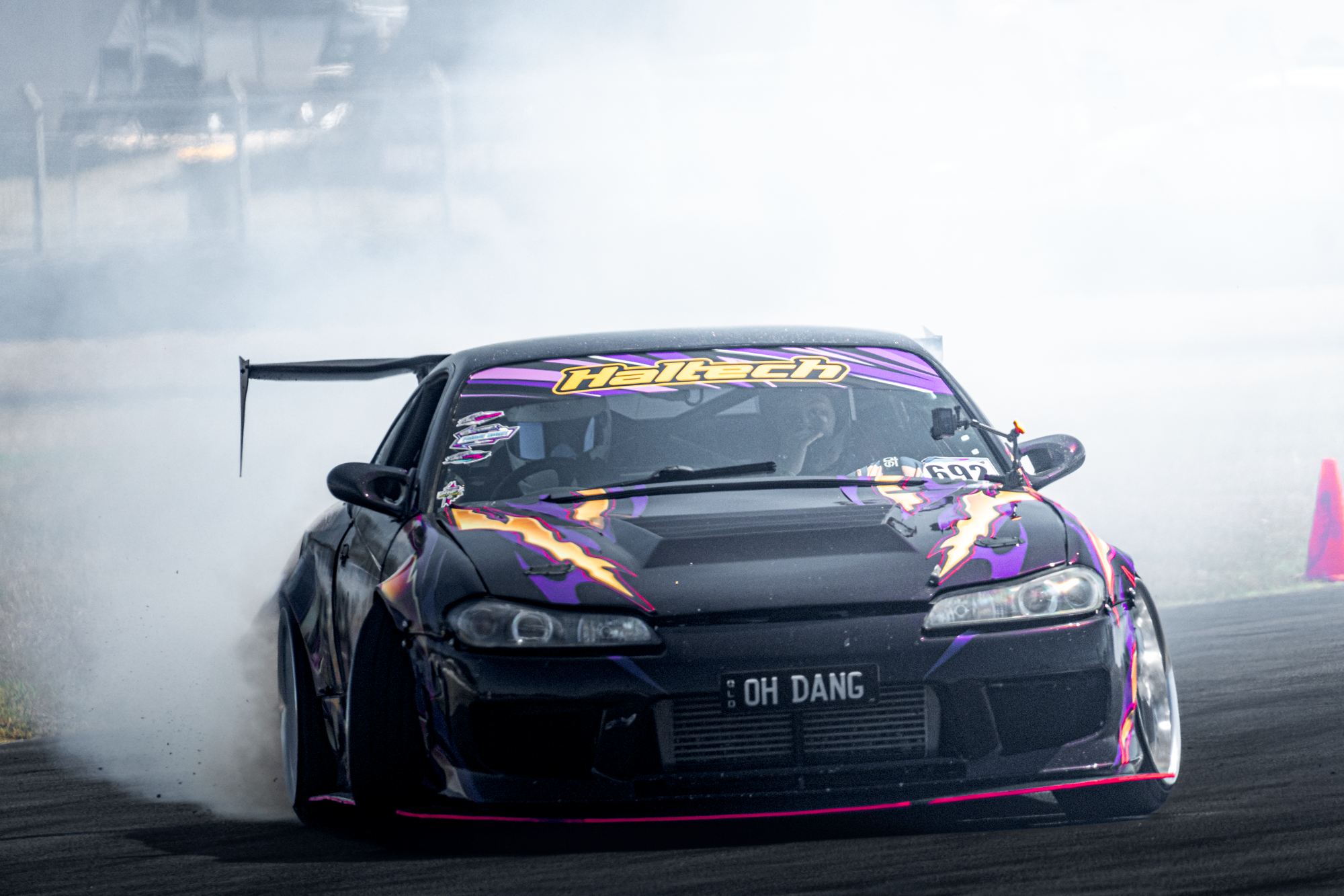 A black sports car with purple, yellow, and pink graphics drifting during a motorsport event, with smoke billowing from the tires.