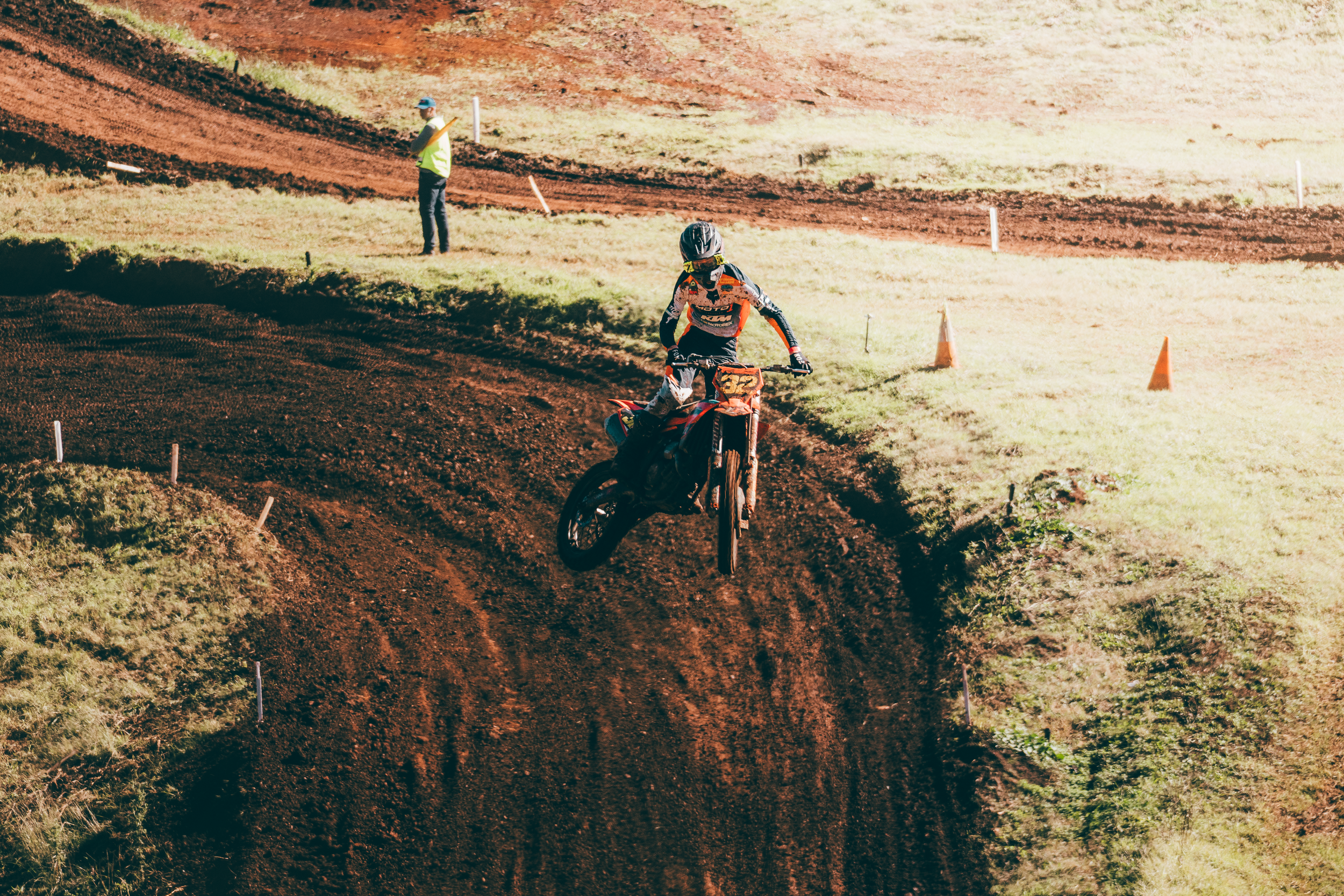 A motocross rider wearing a helmet and racing gear riding on a dirt track with orange cones, with an official standing nearby.