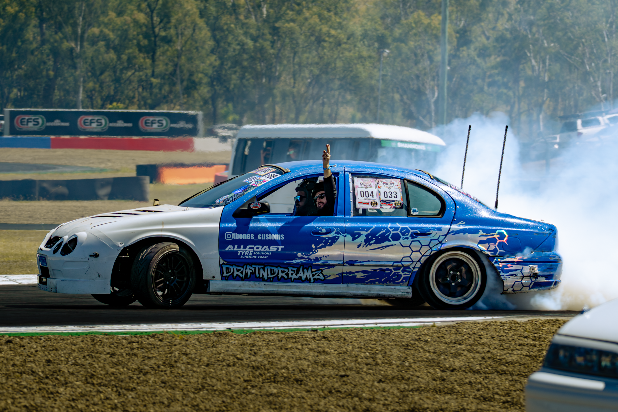 A race car with blue and white design drifting on a track, with smoke billowing behind it. A person inside is raising their hand and making a peace sign. There are banners and trees in the background.