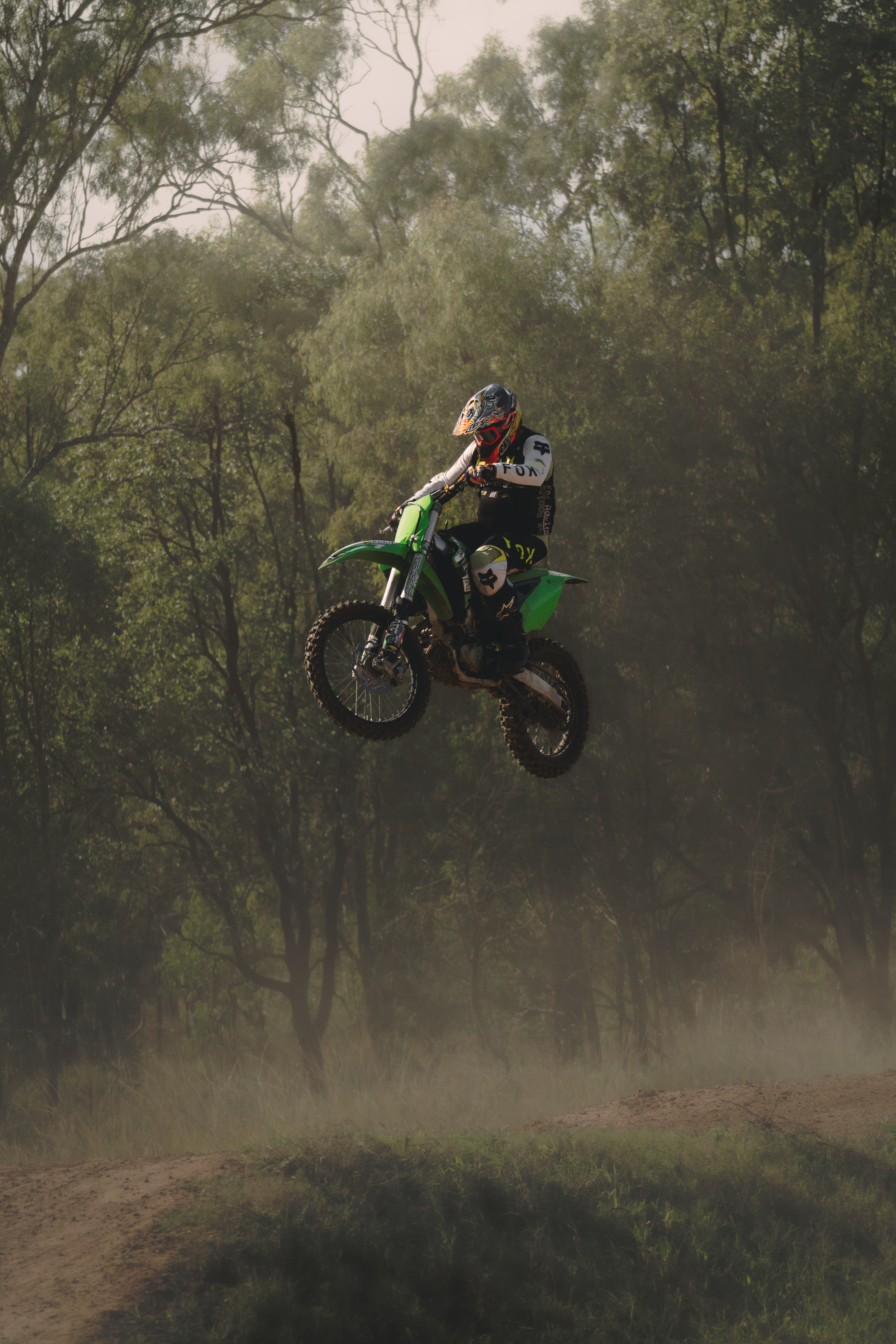 A person wearing a helmet and riding gear performs a jump on a green dirt bike in a wooded area, with trees and dirt trail visible in the background.