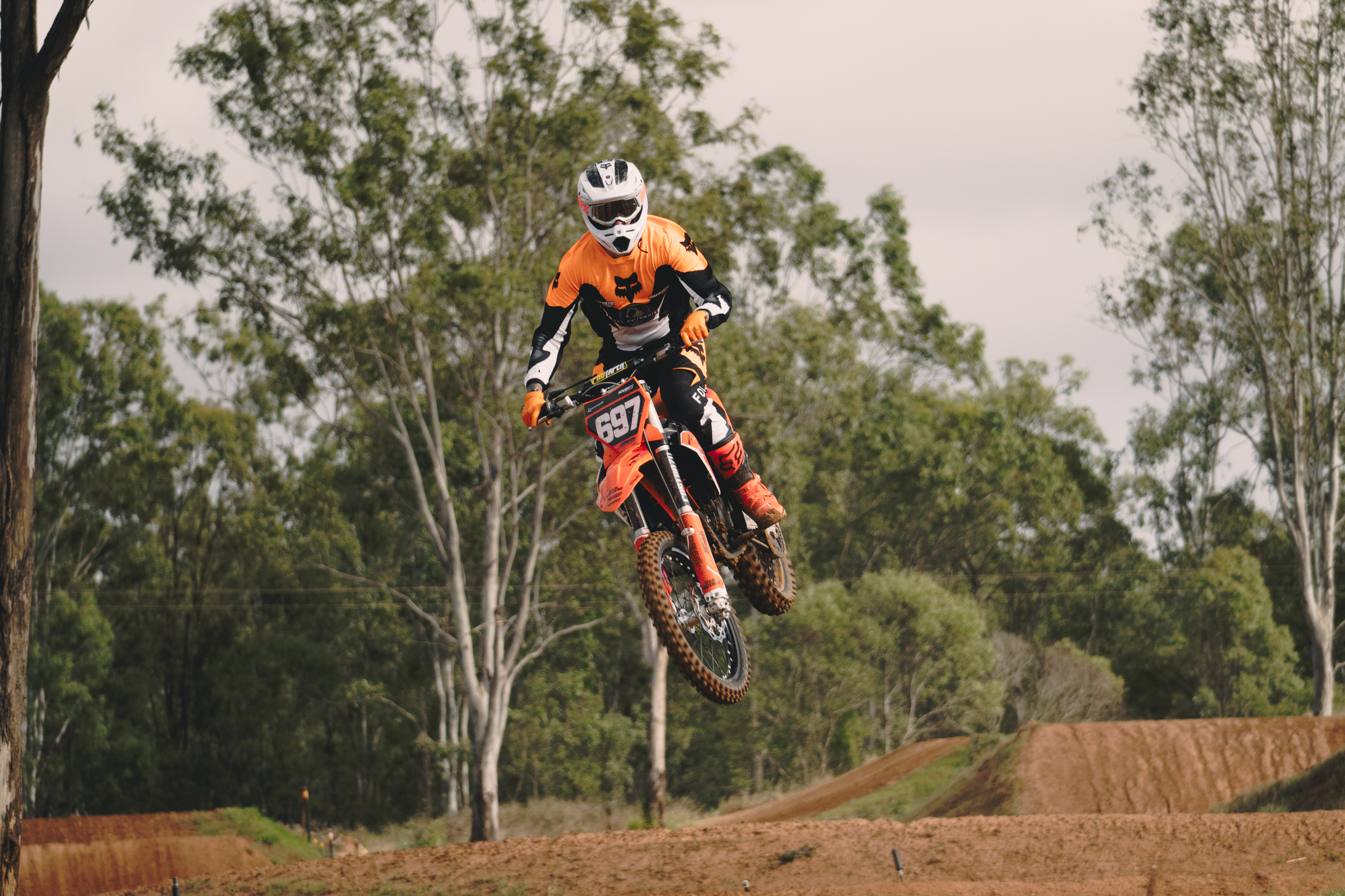 A mountain biker in an orange and black jersey and helmet jumps off a dirt jump on a motocross-style dirt track, with trees and a cloudy sky in the background.