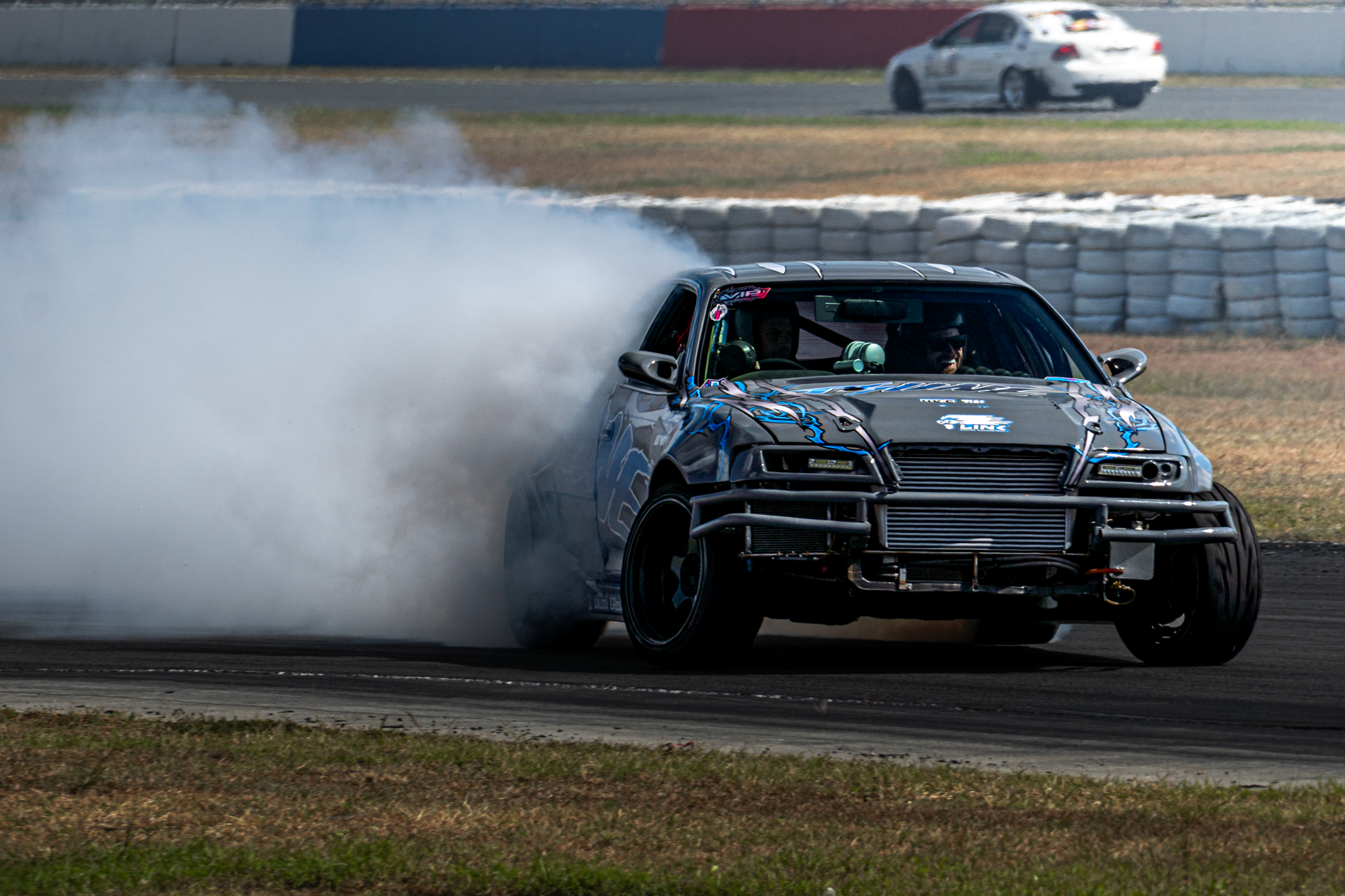 A race car drifting on a racetrack, emitting smoke from its rear tires, with a blurred background of another car and barriers.