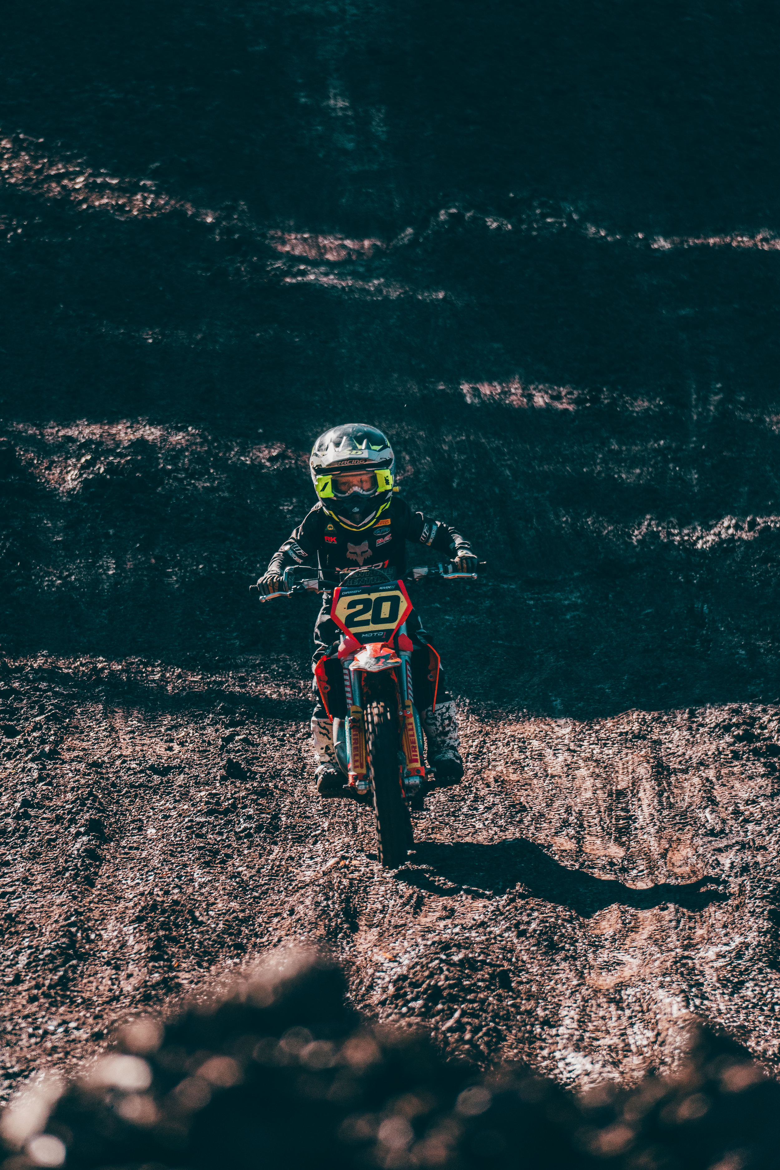 A motocross rider wearing a black helmet and gear, riding a dirt bike on a rough, uneven terrain with the ocean and waves in the background.