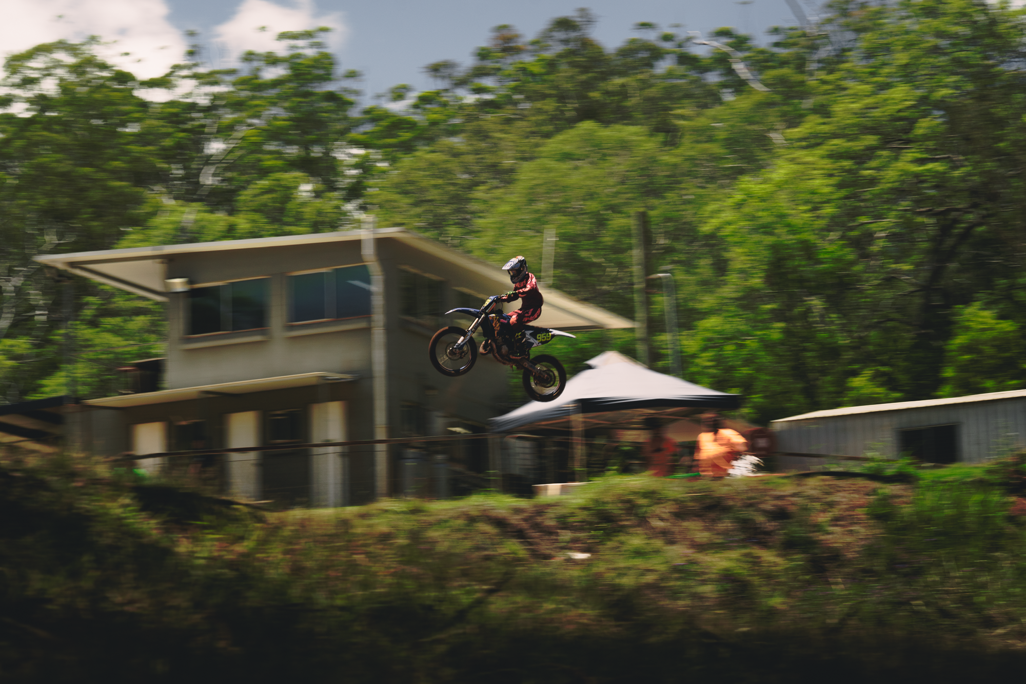 Person riding a motorcycle mid-air during a stunt jump at an outdoor event with spectators and greenery in the background.