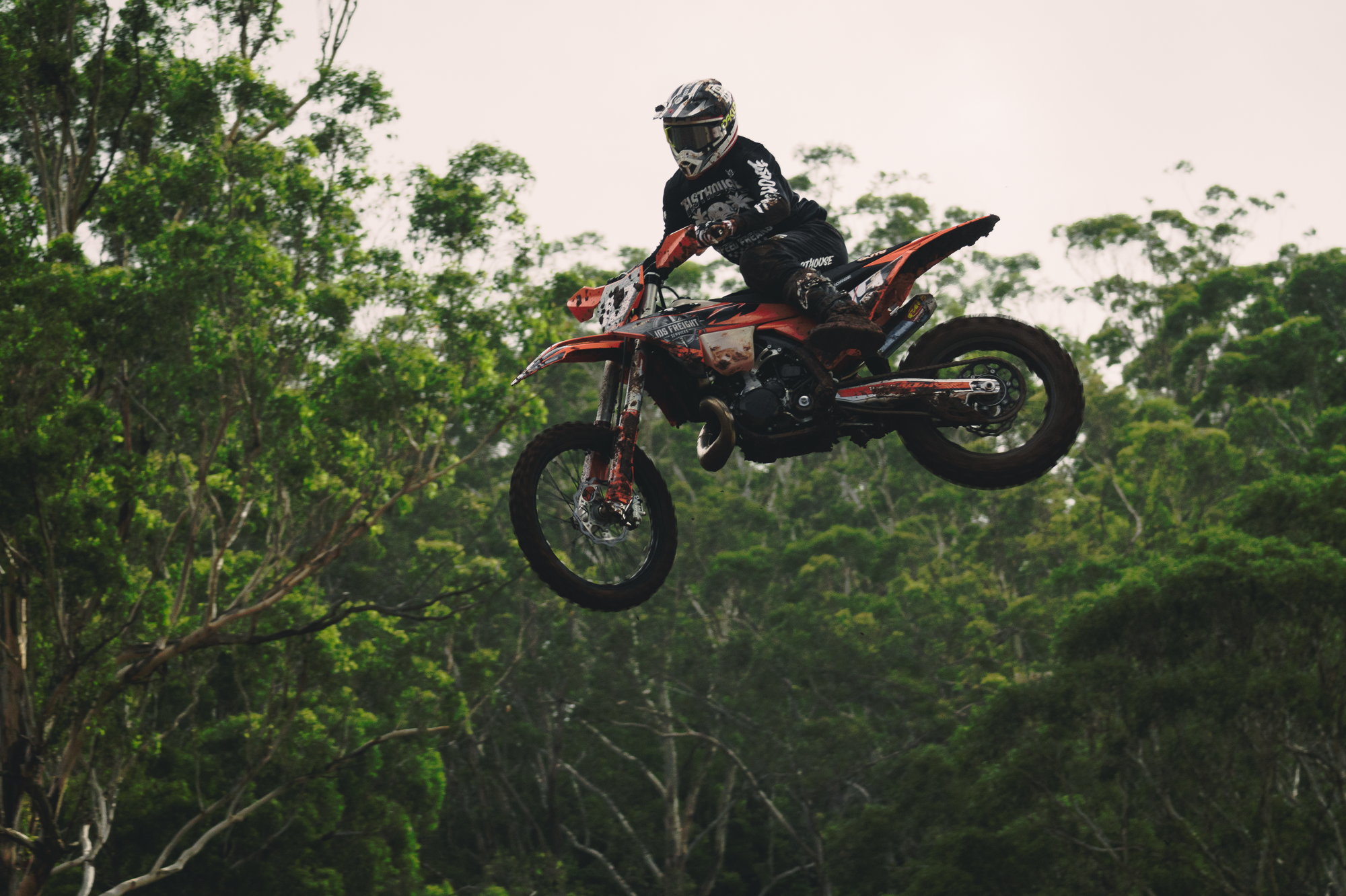 A dirt bike rider mid-air performing a jump in a wooded outdoor area.