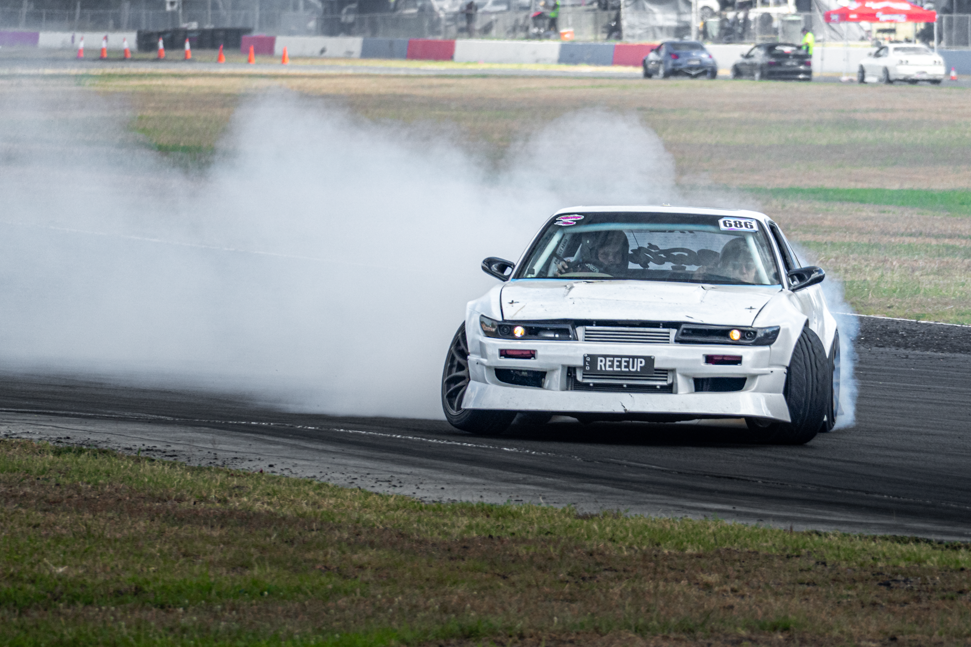 A white race car with the license plate reading 'REEUP' drifting on a dirt track, kicking up a cloud of dust while two other vehicles are parked in the background.