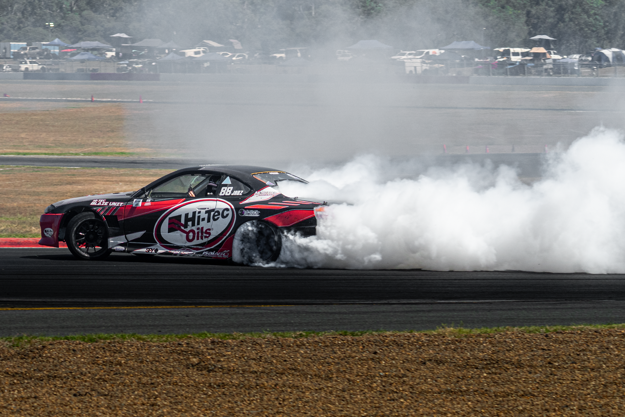 A black and red race car drifting on a track, creating white smoke from the tires with a mist in the air and vehicles and tents in the background.