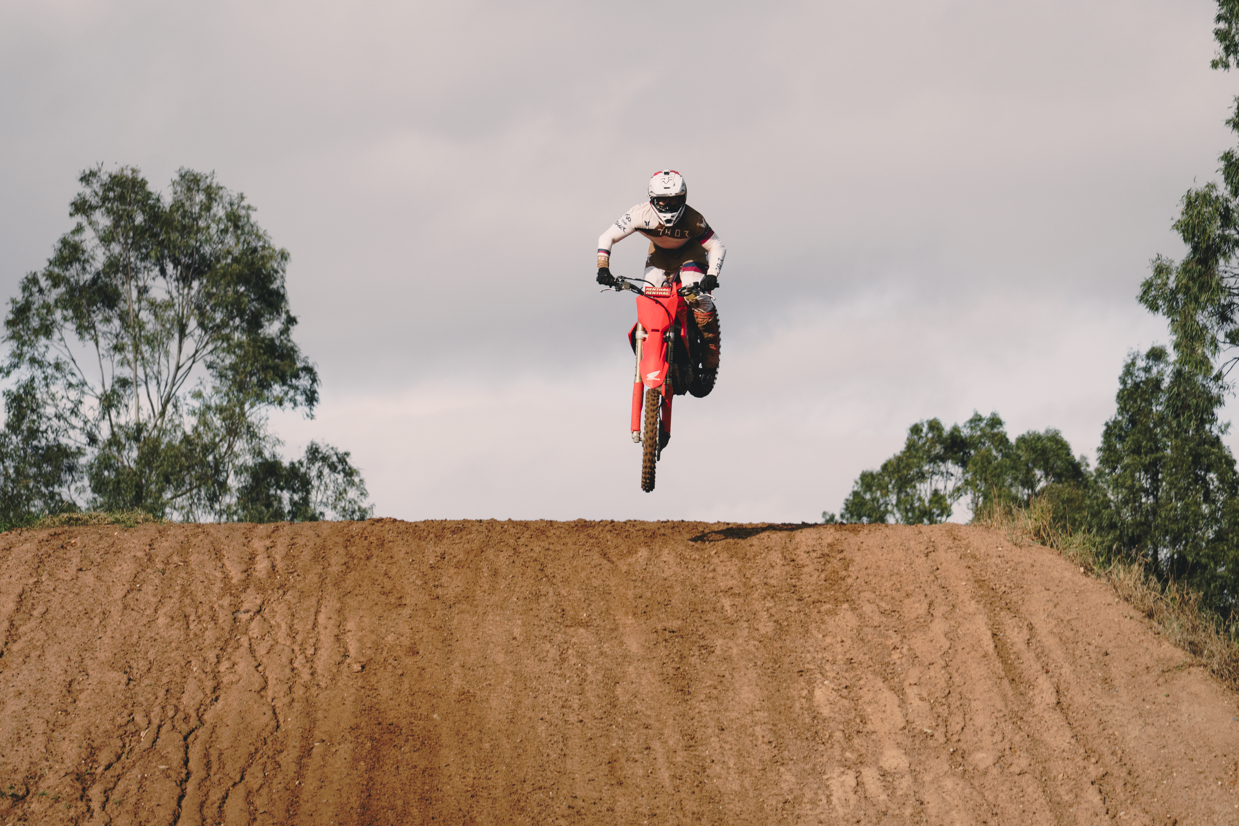 Motocross rider in full gear riding a dirt bike airborne over an inclined dirt track, with tall green trees and a cloudy sky in the background.