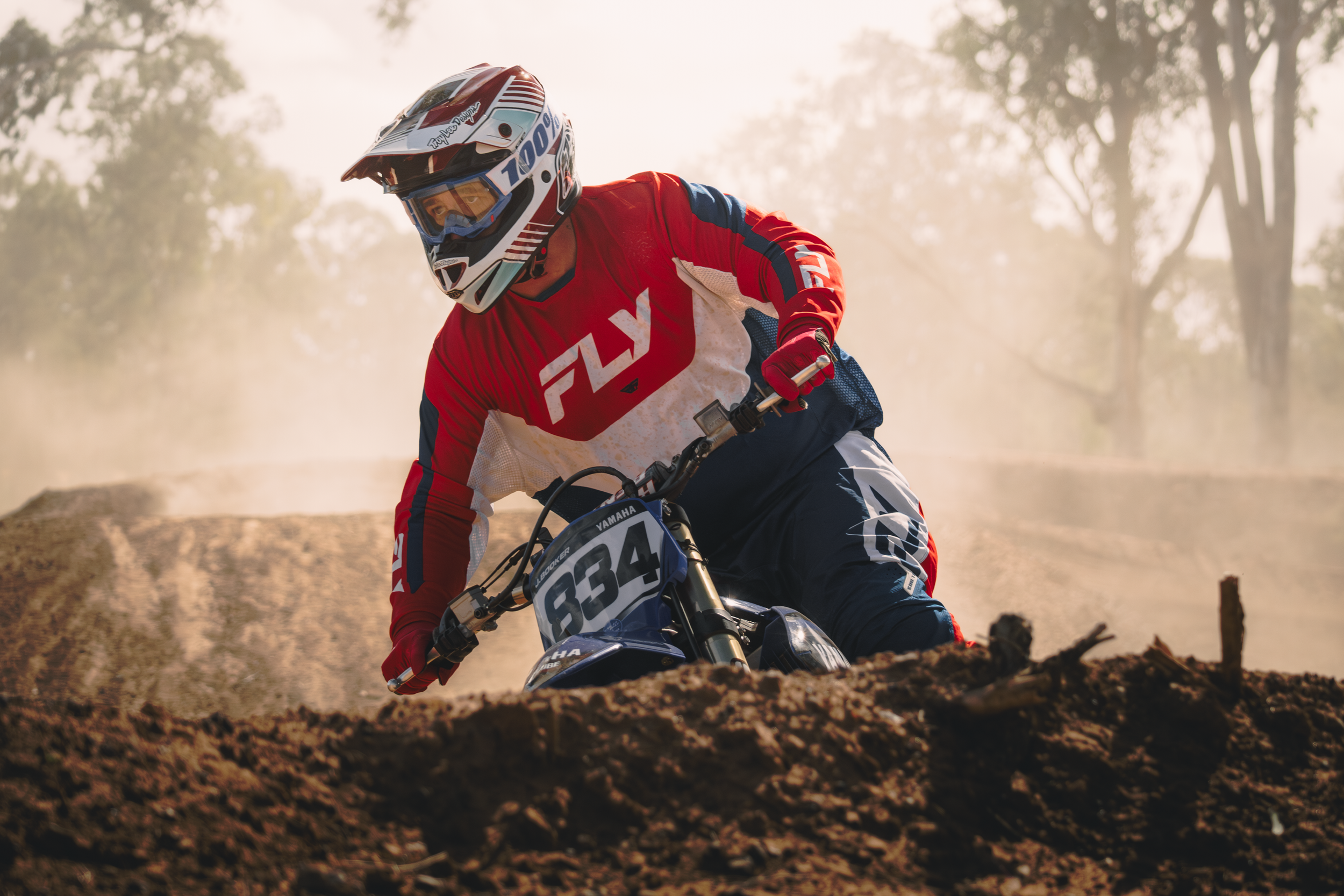 A motocross rider in a red and blue racing suit with a helmet and goggles, riding a Yamaha dirt bike on a dirt track with dust and trees in the background.