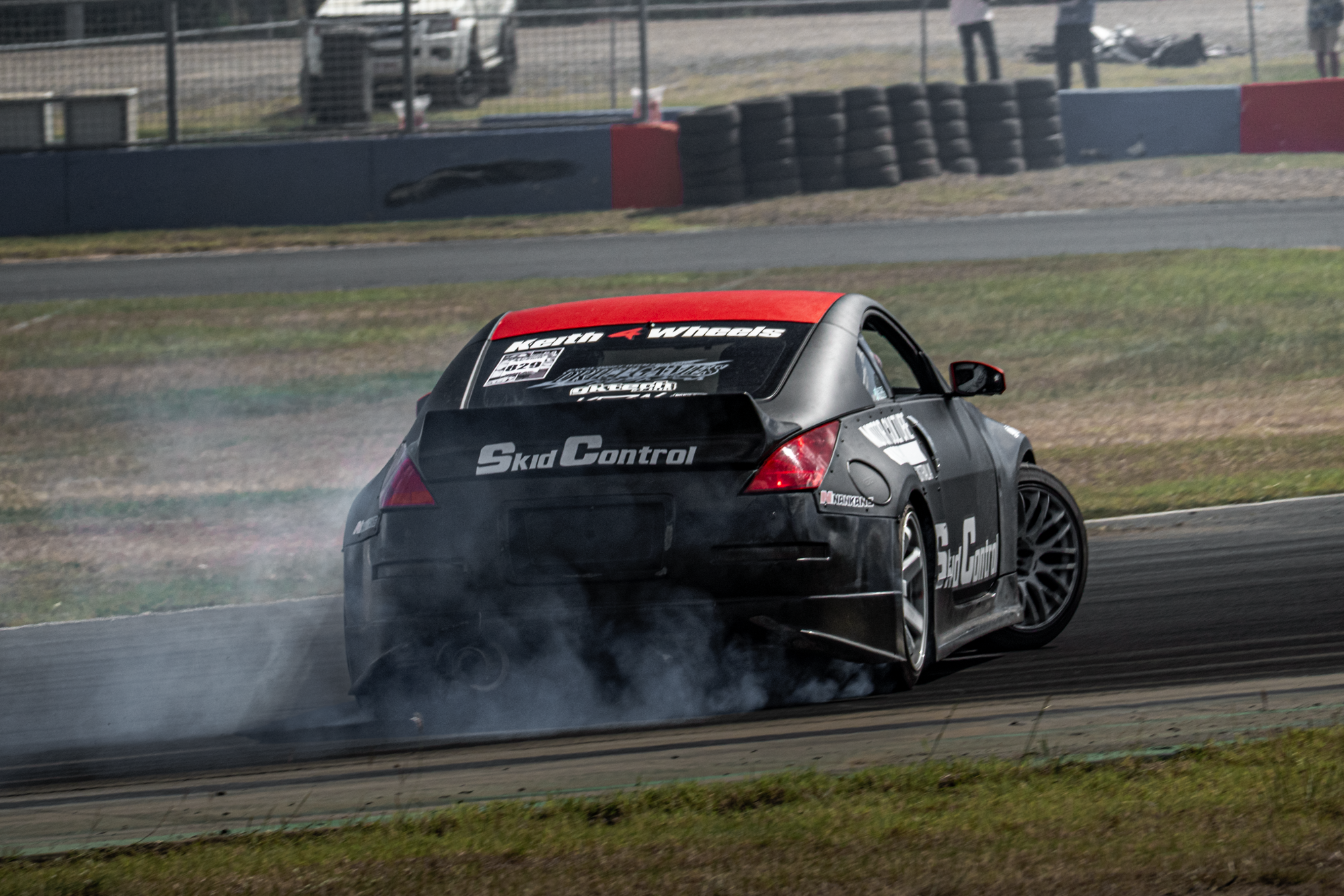 A black race car with red accents drifting around a track, emitting smoke from its tires with a fence and tire barriers in the background.