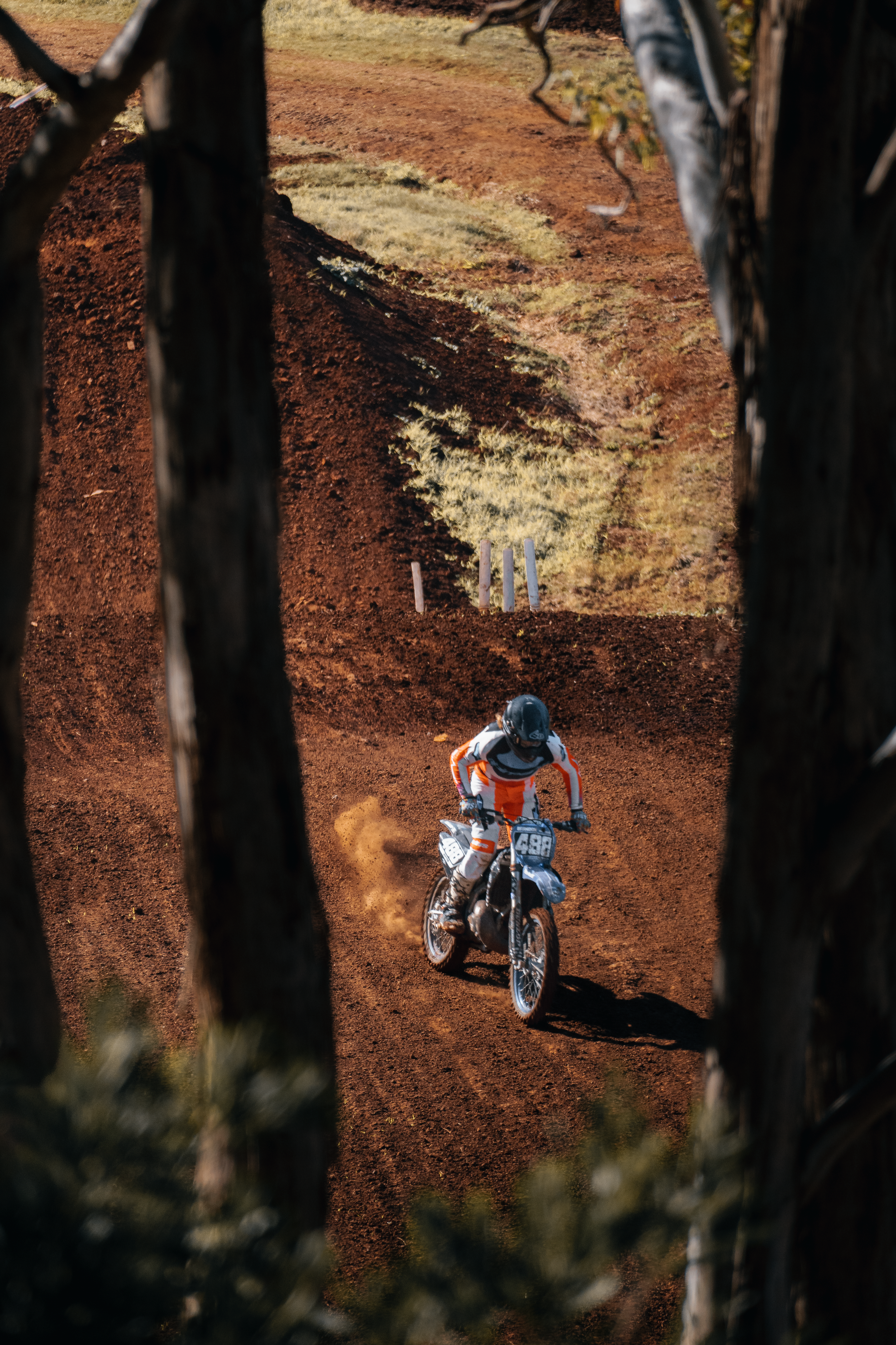 A motocross rider in full gear riding on a dirt track seen through a gap in trees.