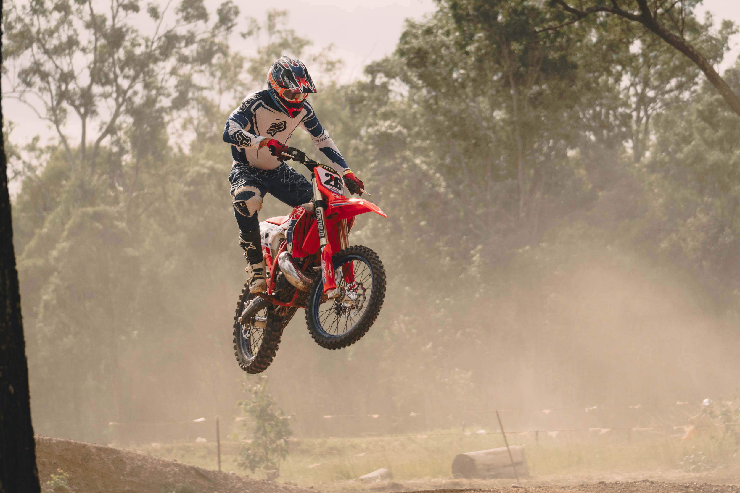 A motocross rider wearing a helmet and racing gear riding a red dirt bike mid-air on a dirt track surrounded by trees.