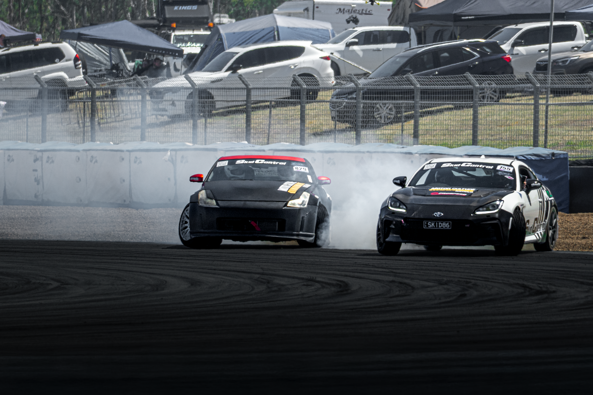 Two race cars drifting on a track, creating smoke behind them, with a background of parked vehicles and tents.