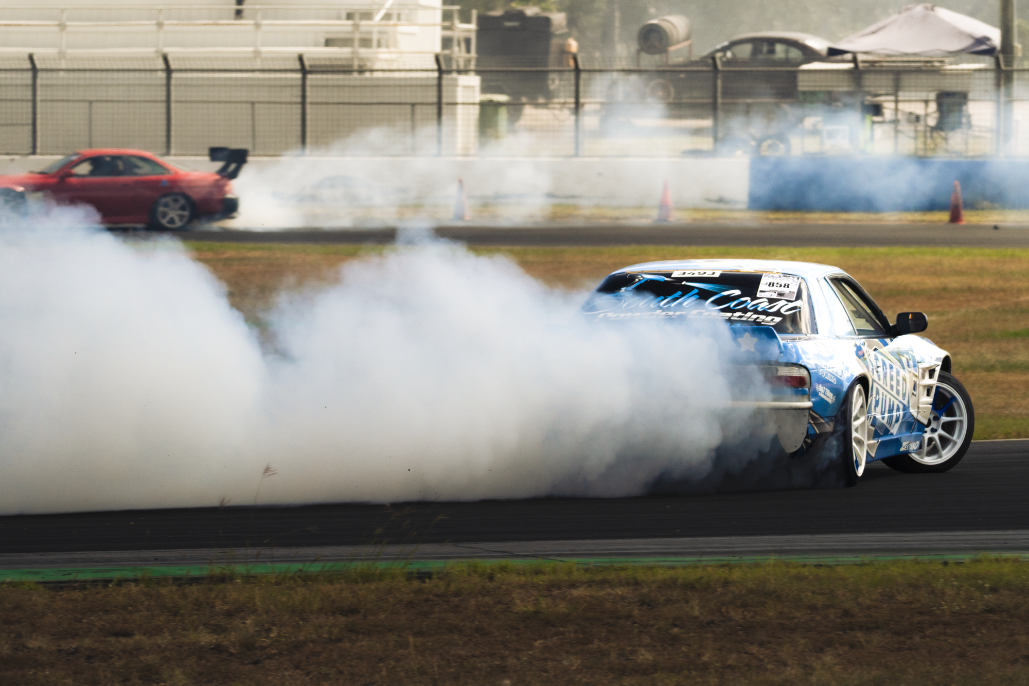 A blue race car drifting around a corner on a racetrack, creating thick white smoke from its tires. A red car is visible in the background, along with safety barriers and orange cones on the track.