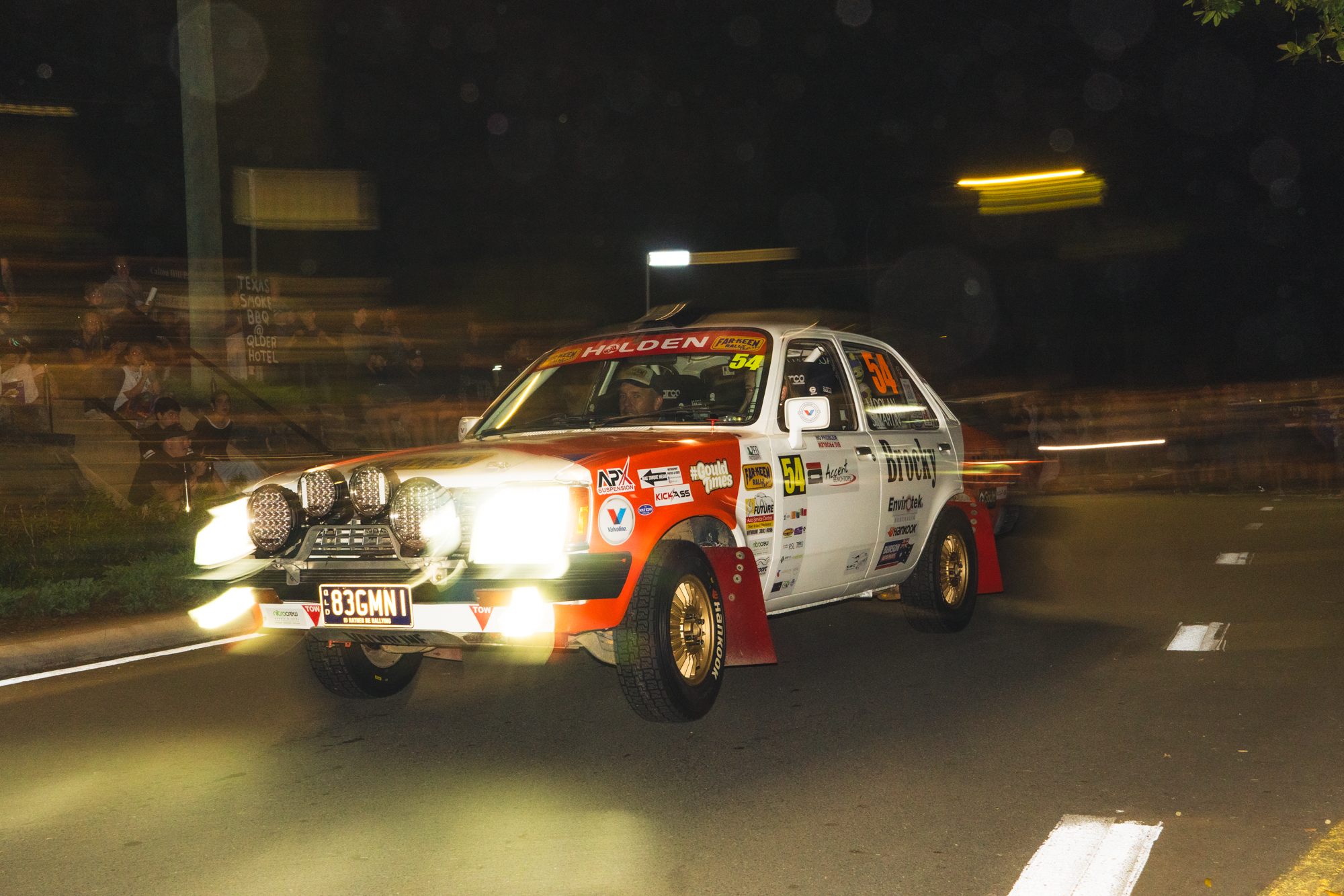 A rally car with multiple sponsor stickers and bright headlights driving on a dark road at night, with blurred lights and spectators in the background.