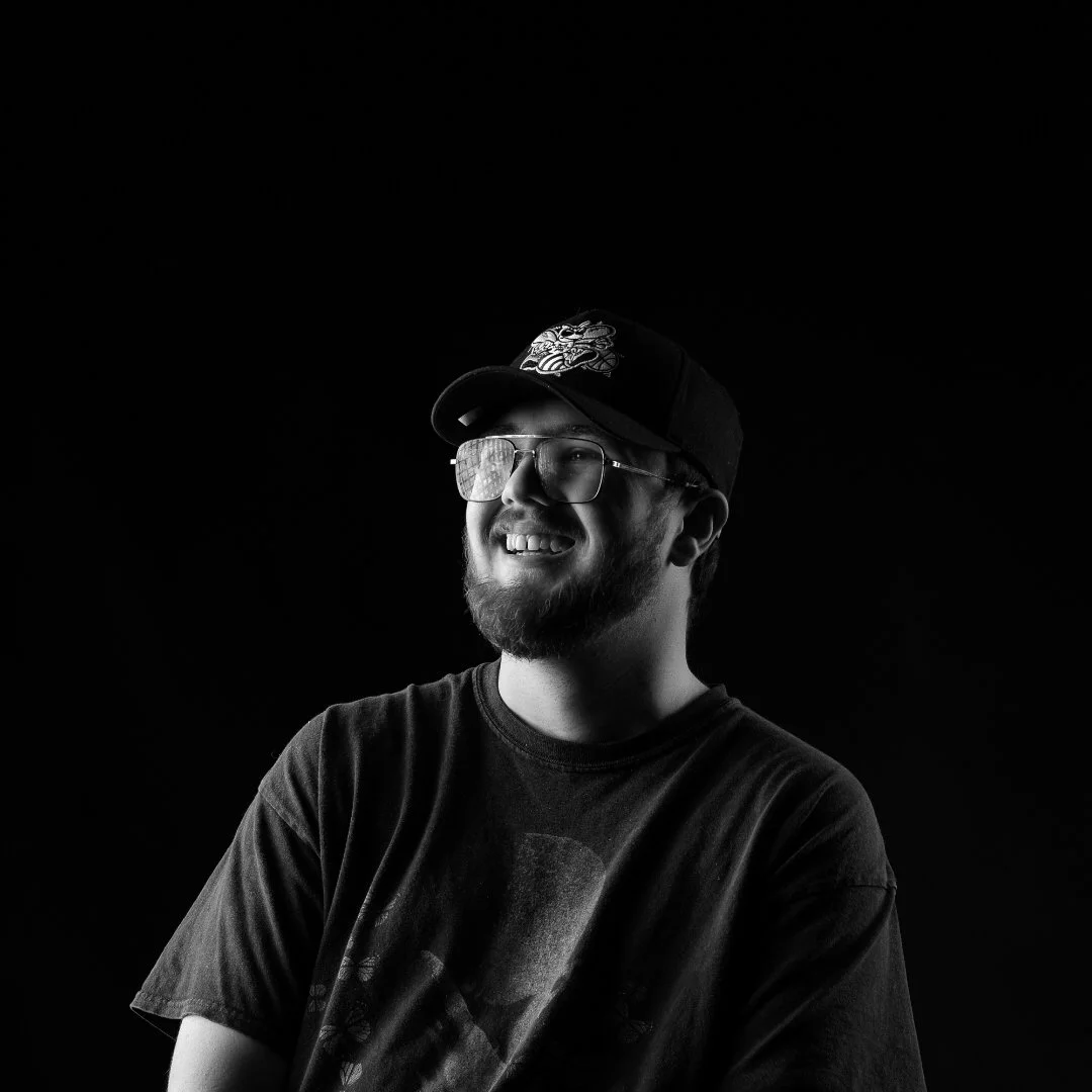 Black and white portrait of a smiling man wearing glasses, a baseball cap with a graphic, and a T-shirt, against a dark background.