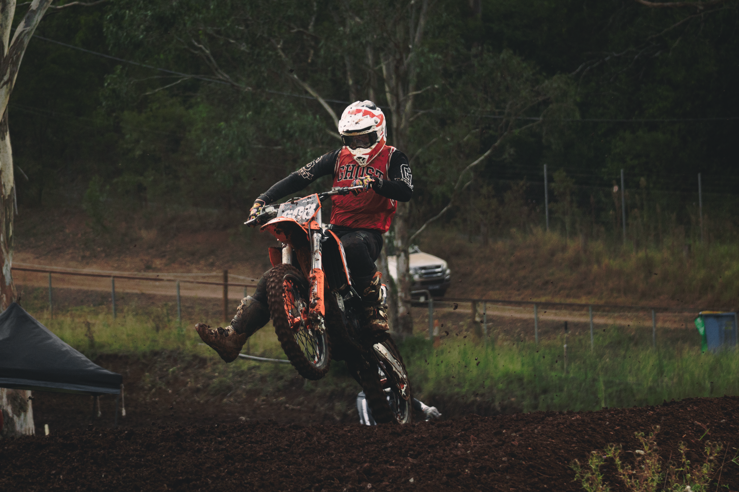 A motocross rider wearing a helmet and protective gear jumps over a dirt mound on a motocross track surrounded by trees.