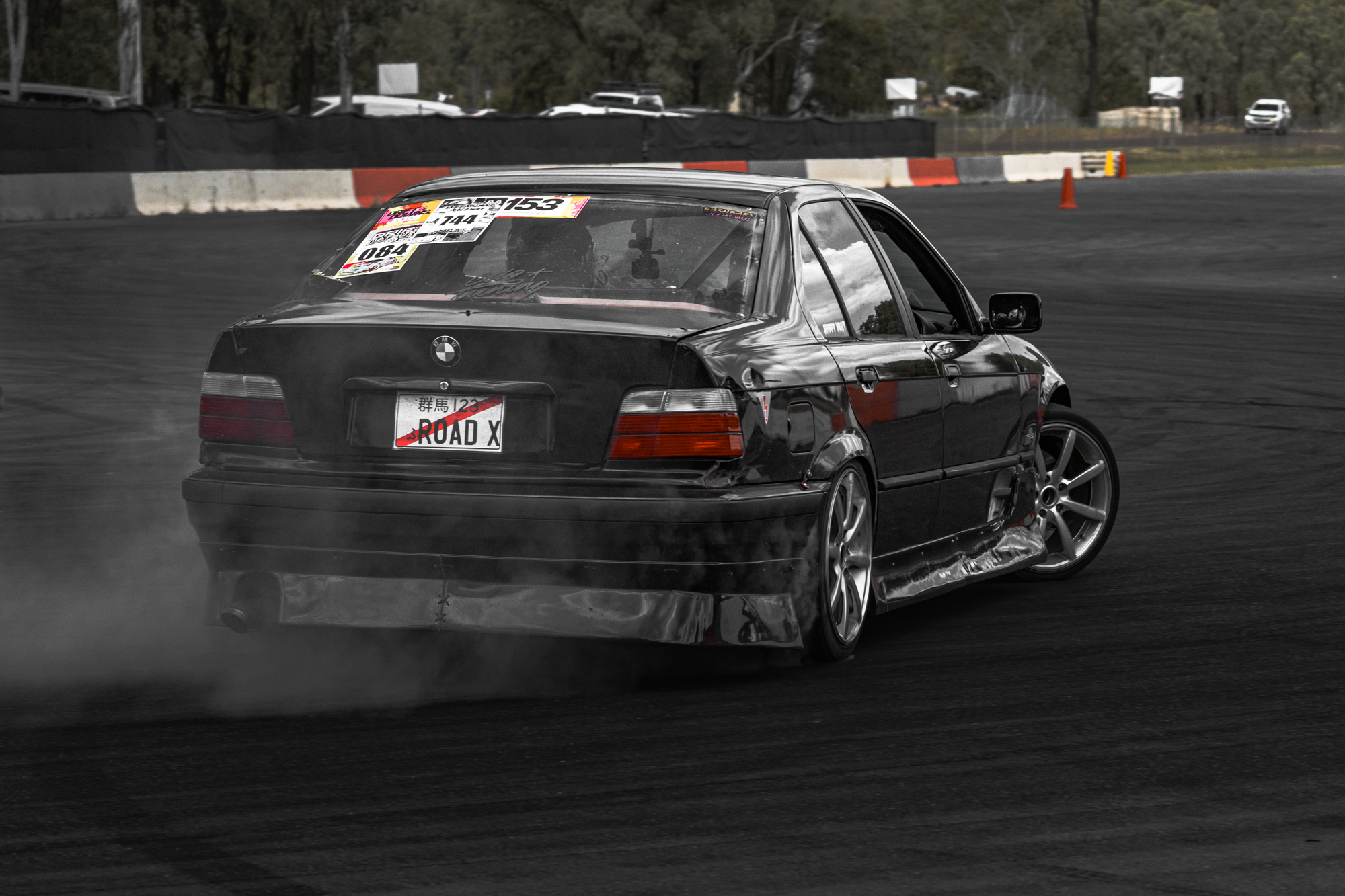 A black BMW car drifting on a race track, creating smoke from its tires with a scenic background of trees and parked vehicles.