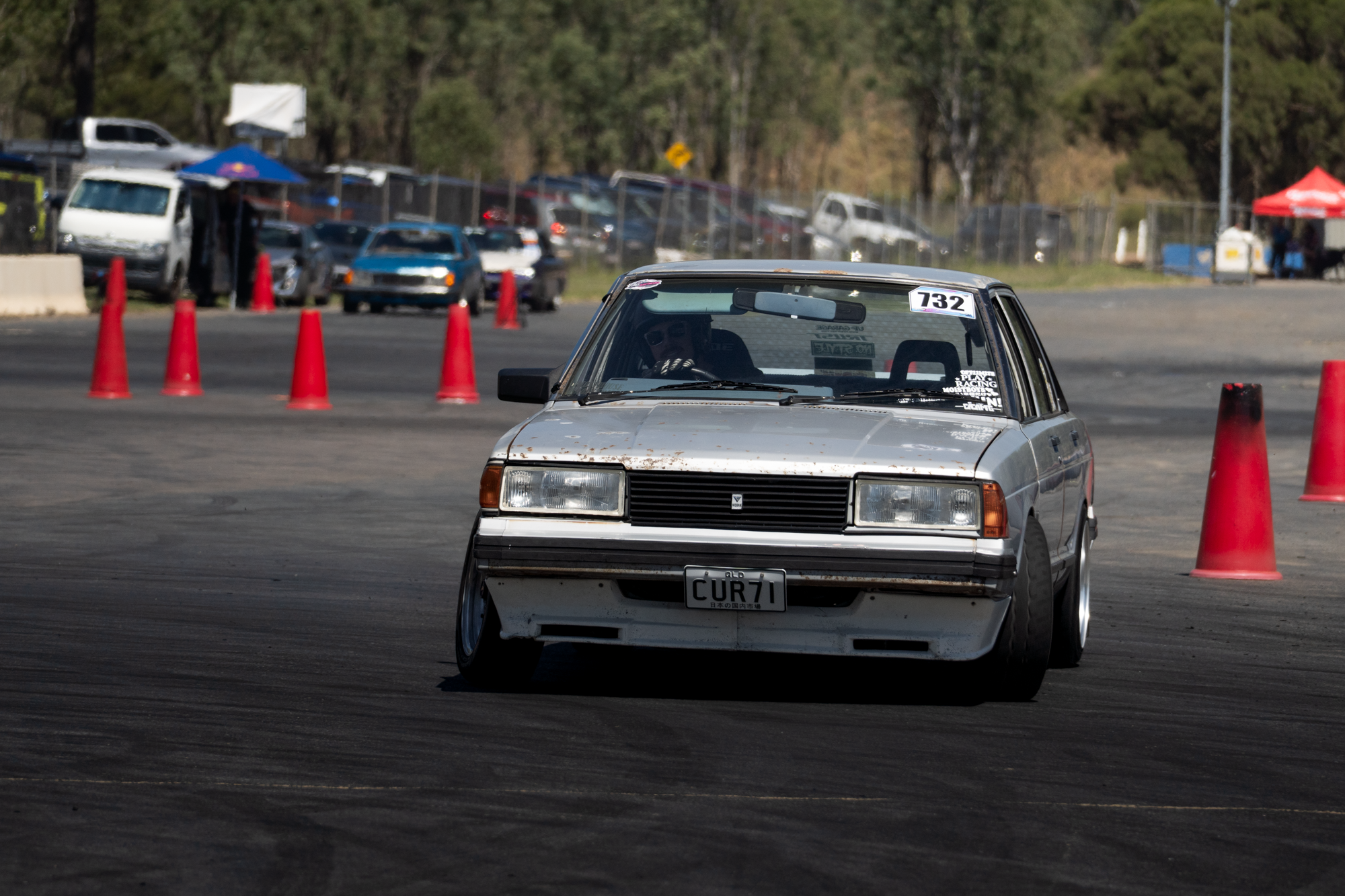 Vintage silver sedan with license plate 'CUR71' navigating an outdoor autocross course marked by orange traffic cones. In the background, there are several parked and moving cars, a tent, and a forested area.