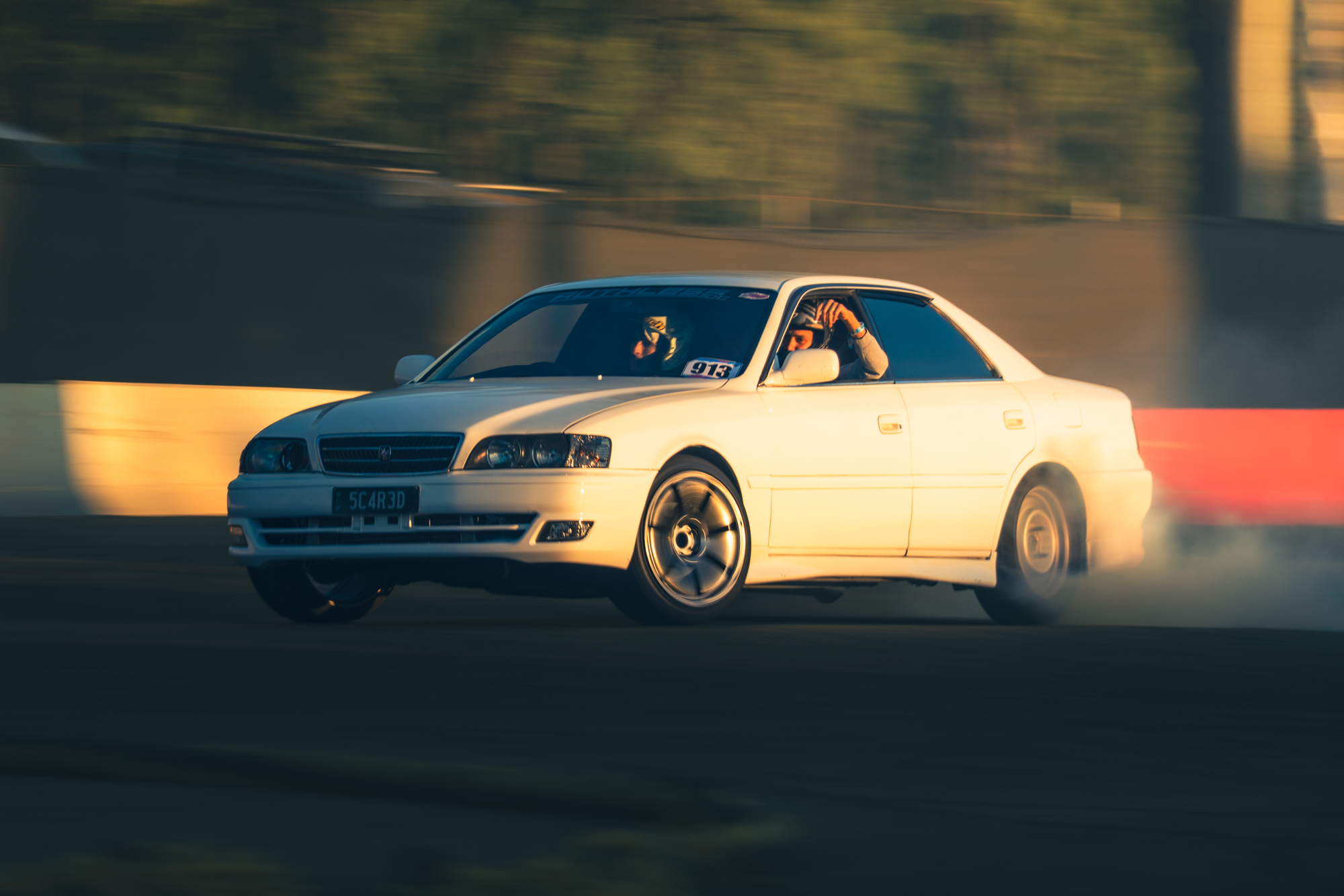 A white car drifting on a race track with smoke coming from its rear tires, driver is visible wearing a helmet, motion blur in background indicating speed.