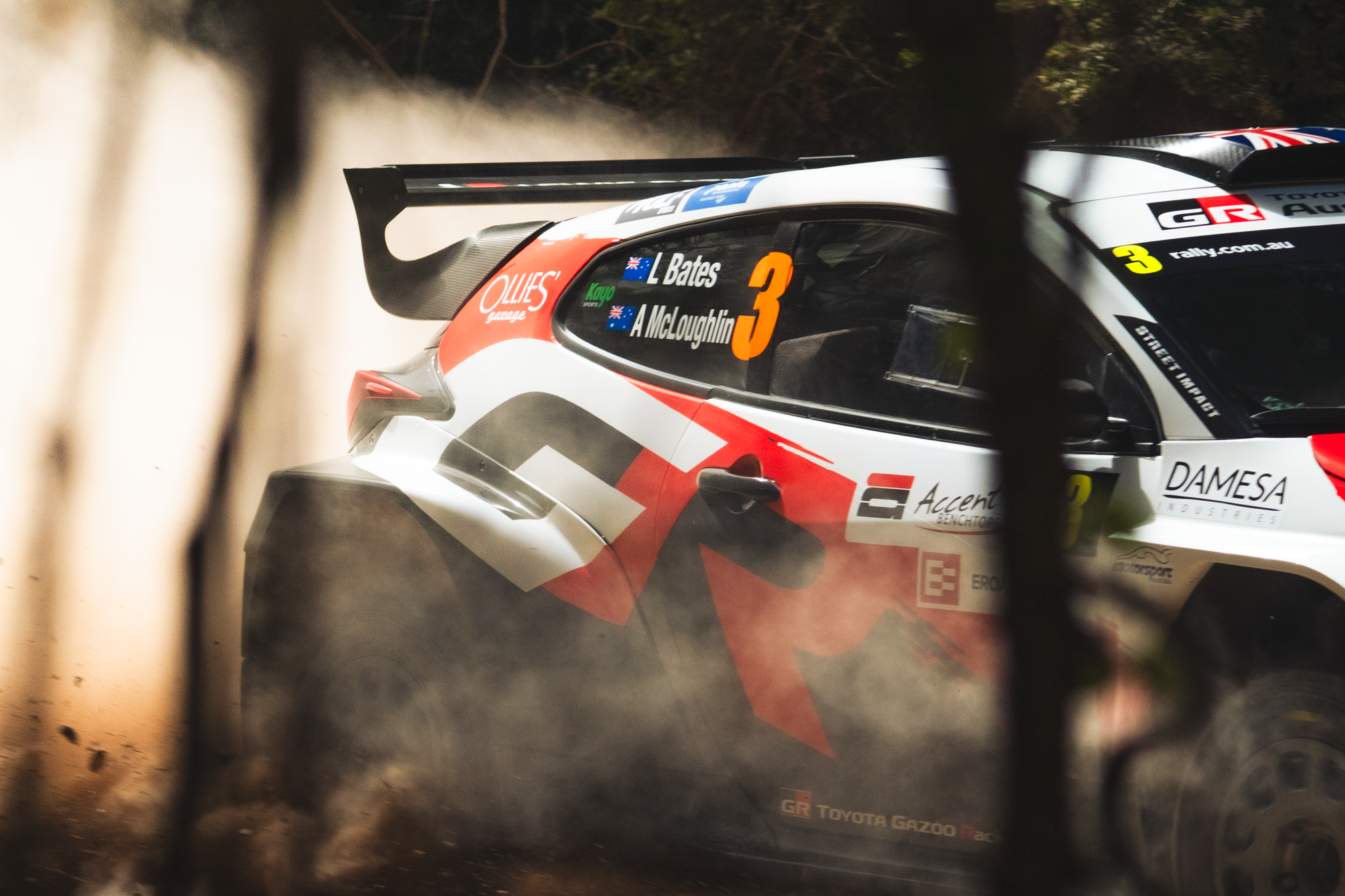 A rally car driving on a dirt road, kicking up dust, with the driver’s side window showing the names L. Bates and A. McLoughlin and various sponsor logos.