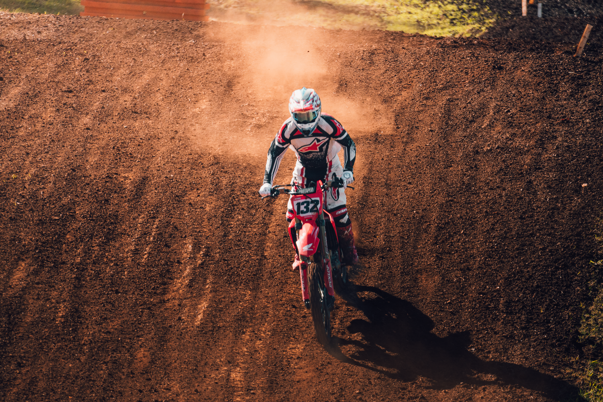 A person in motocross gear riding a red dirt bike on a dirt track, kicking up dust with a background of a dirt hill and greenery.