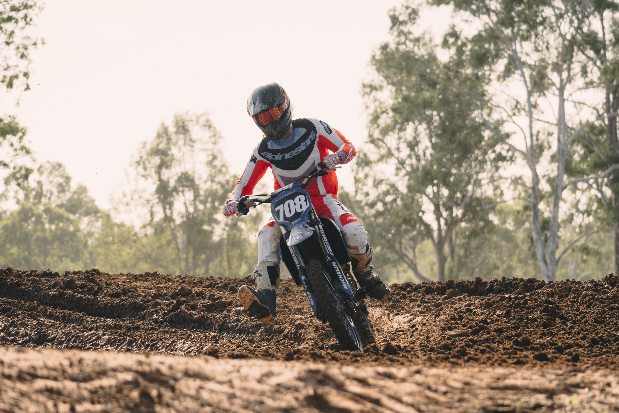 A motocross rider wearing a helmet and colorful gear riding a dirt bike on a rough, muddy track with trees in the background.