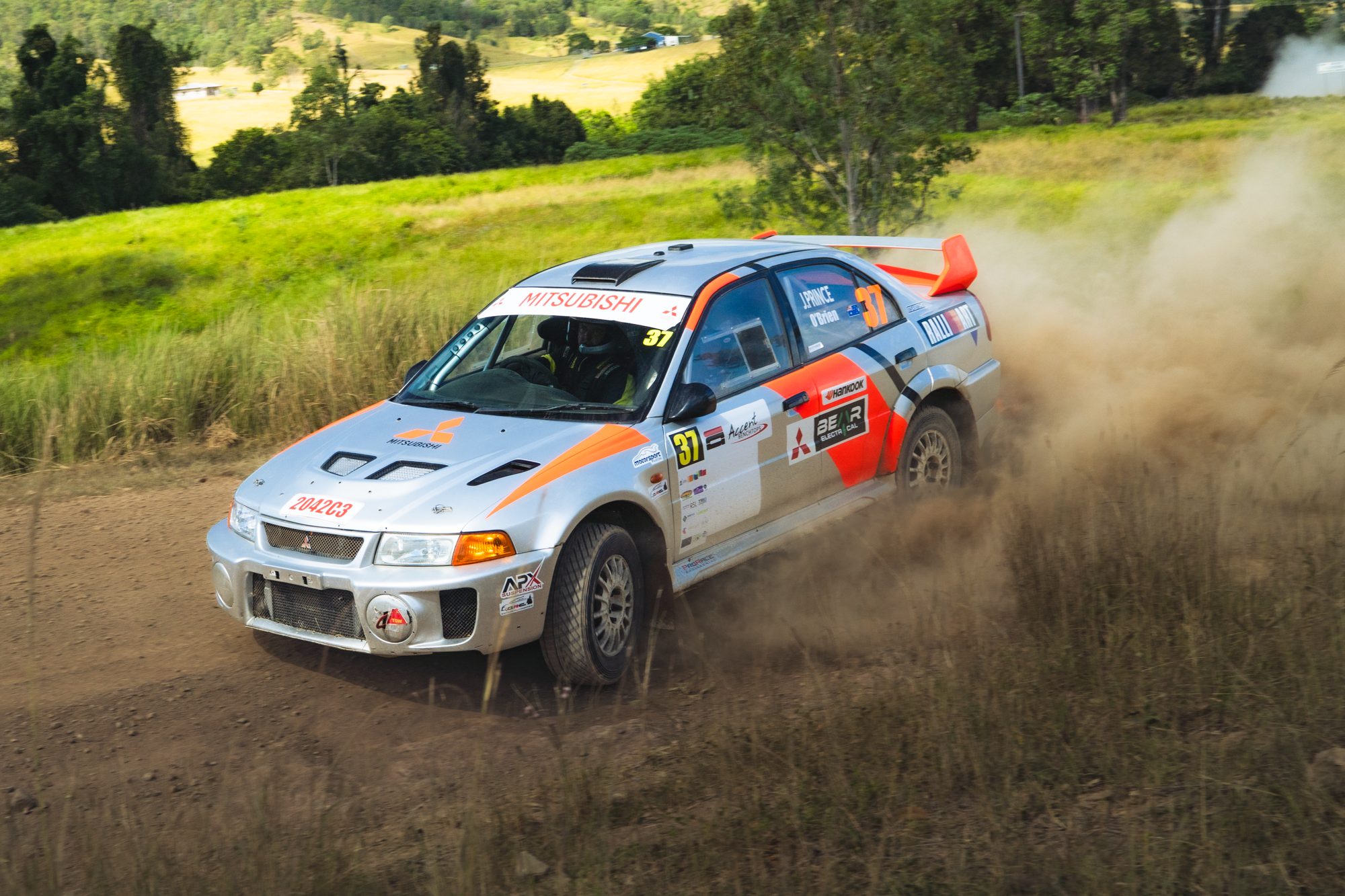 A silver rally car with orange accents speeding on a dirt road, kicking up dust in a rural area with green trees and fields in the background.