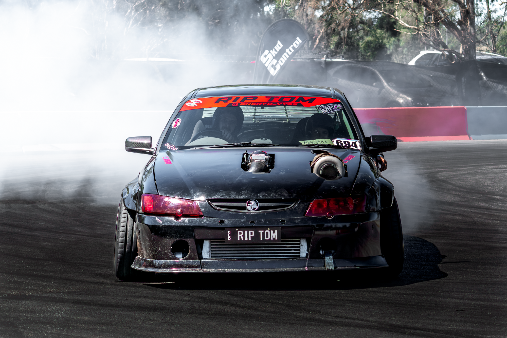A black sports car with a prominent front-mounted engine, drifting on a race track with smoke billowing behind it. The car has a custom license plate that reads 'RIP TOM' and various decals, including a red banner on the windshield with the text 'RIP