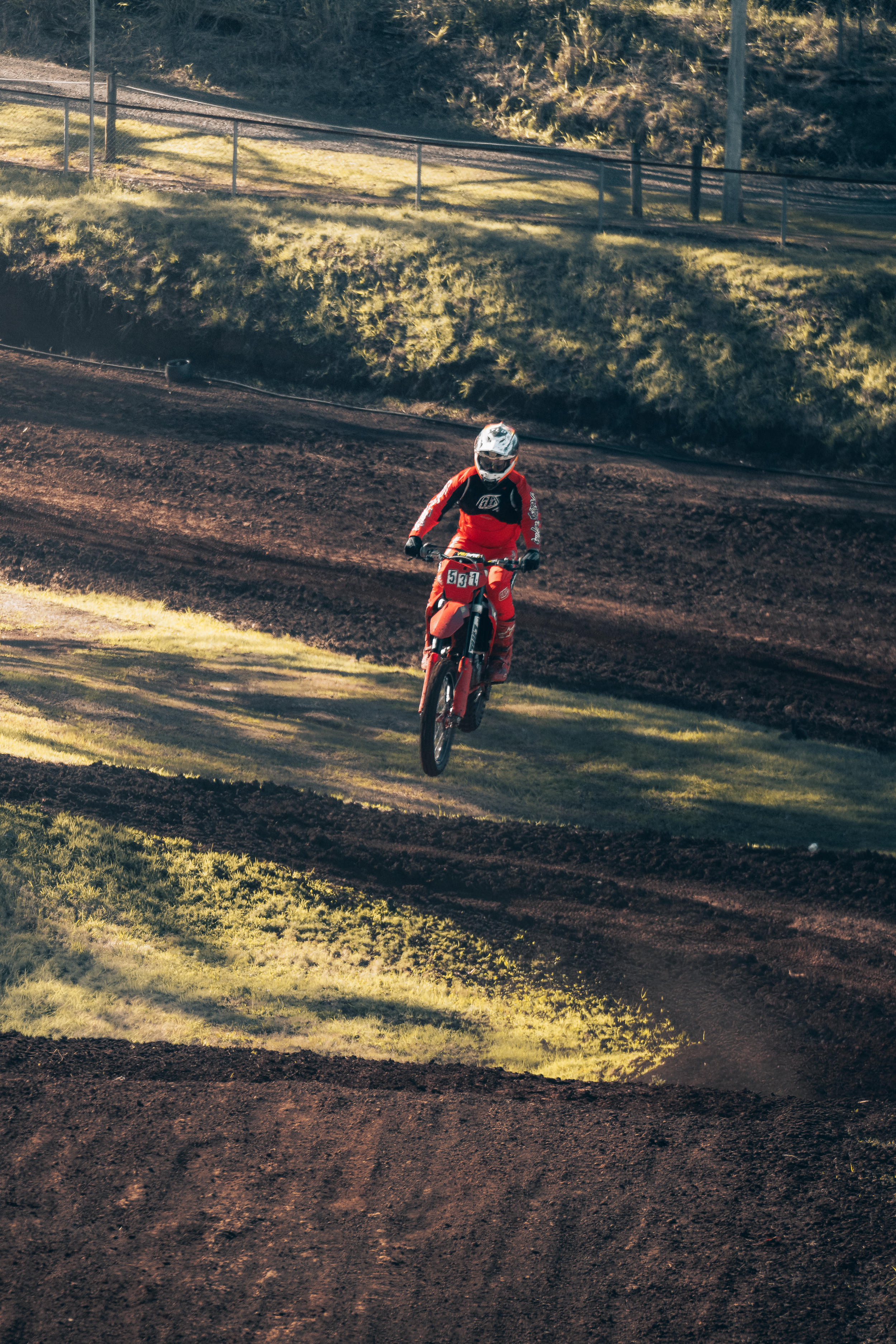 A person riding a dirt bike on a motocross track during daytime, with grass and trees in the background.