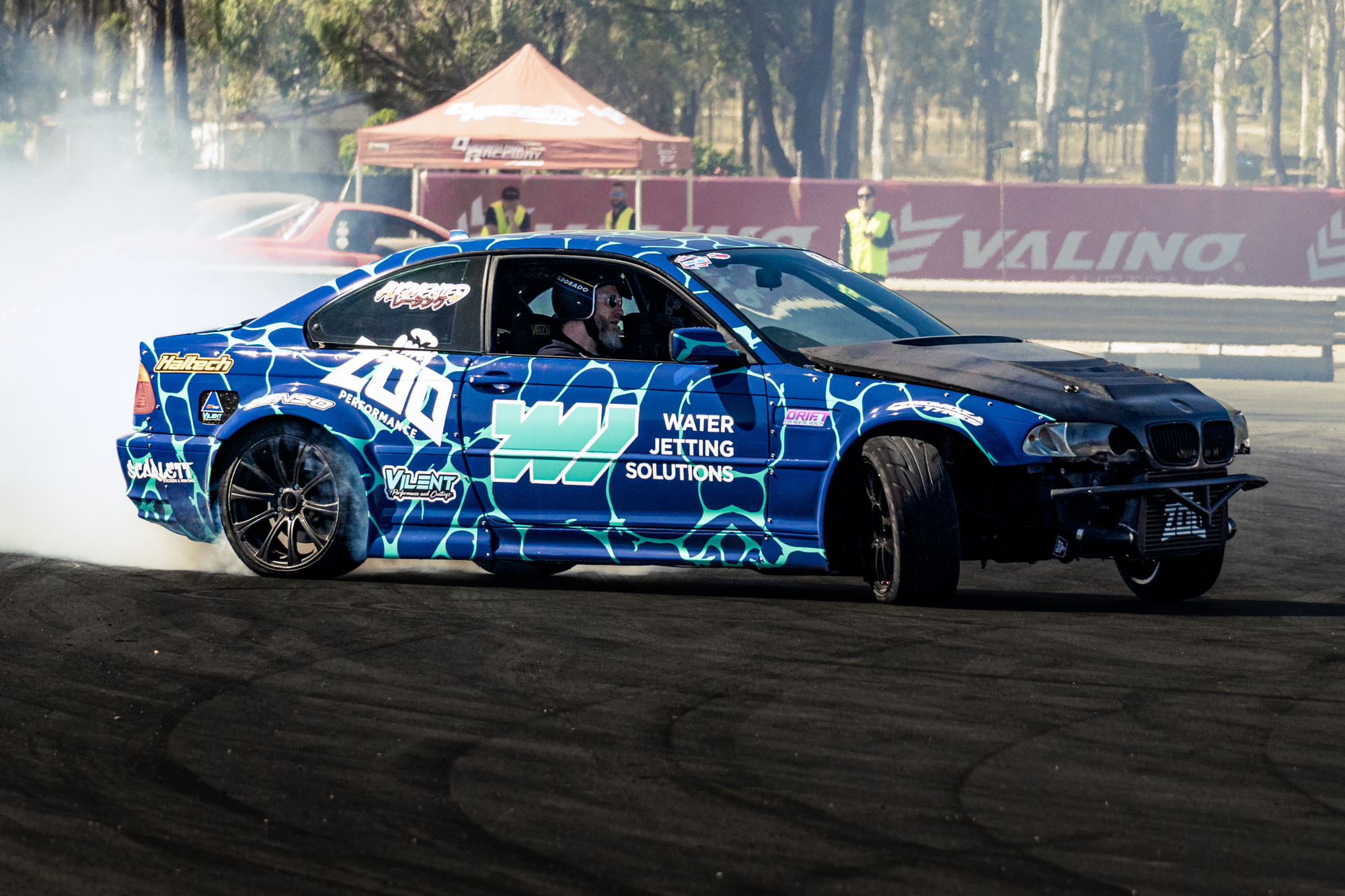A blue modified BMW car performing a drift on a race track, emitting smoke from the rear tires, with a driver wearing sunglasses and a helmet. In the background, there are orange tents, a red car, and track officials in yellow vests.