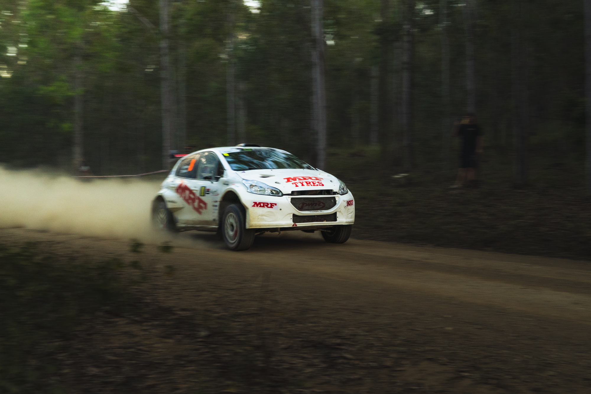A white rally car speeding on a dirt track through a wooded area, kicking up dust behind it, with a person standing in the woods to the right observing the race.