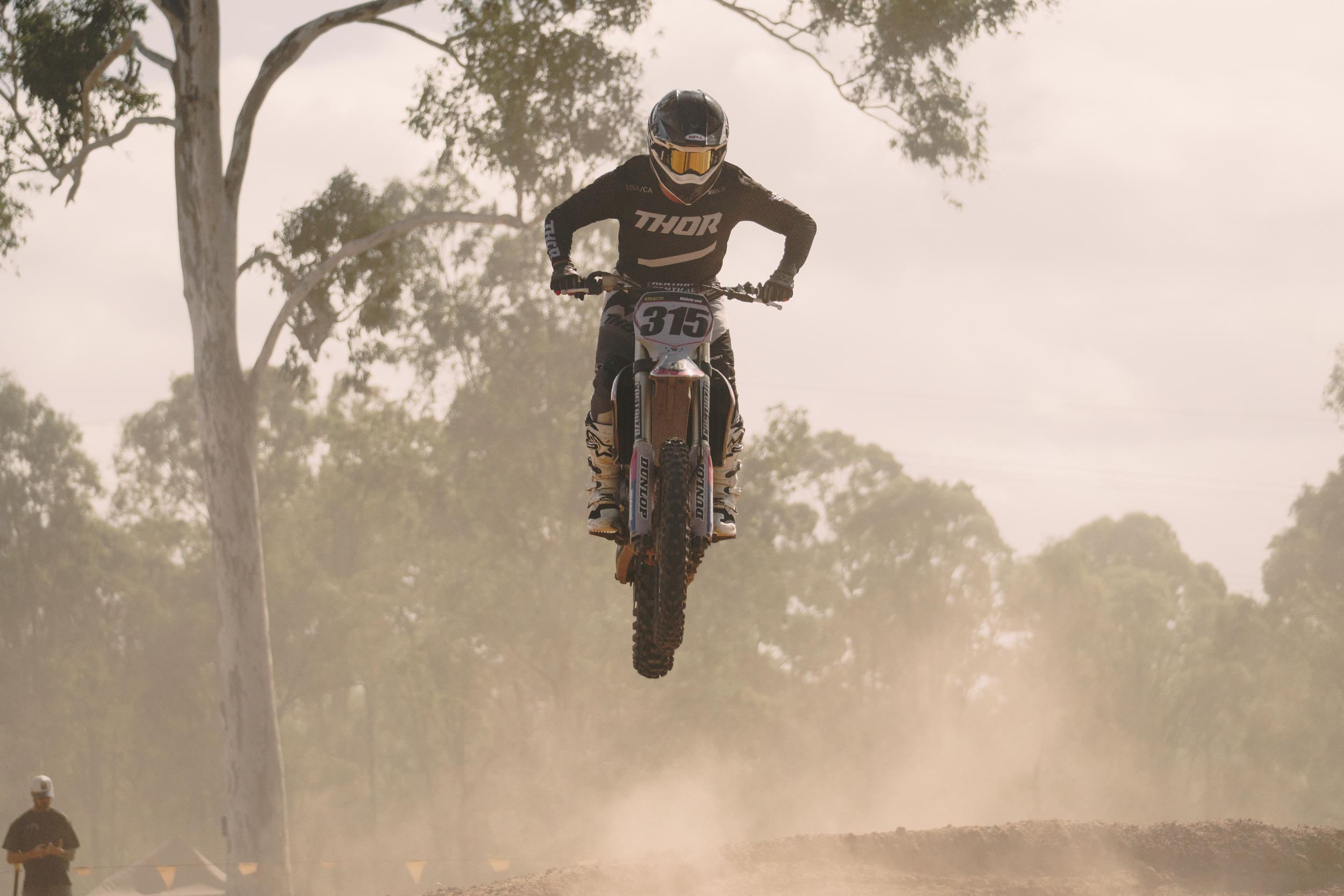 A mountain biker wearing a black helmet and black gear with 'THOR' branding, riding a dirt bike in mid-air over a dirt track with trees in the background and dust clouds behind.