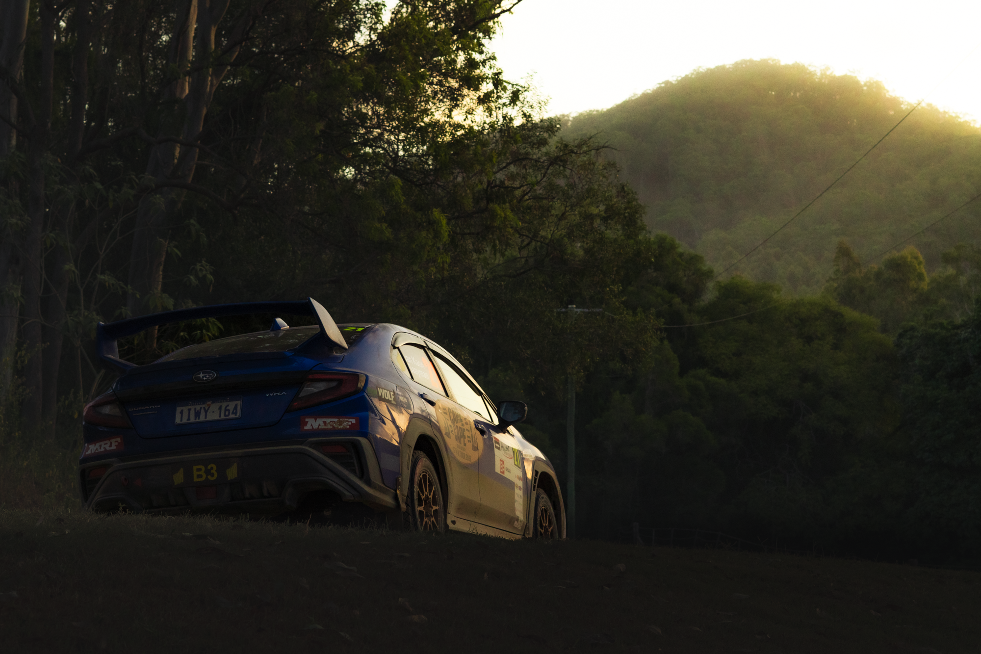 A racing car with various sponsor stickers parked on a grassy area during sunset, surrounded by trees and hills in the background.