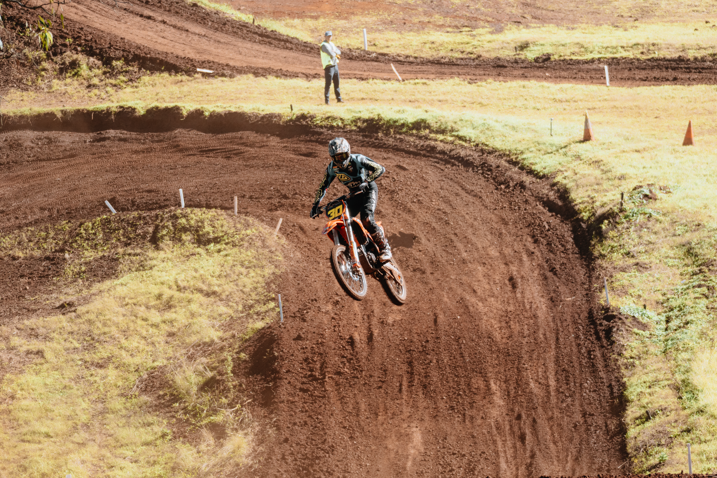 A dirt bike rider wearing a helmet and riding gear, navigating a motocross trail with dirt mounds and grassy areas. An observer stands in the background.